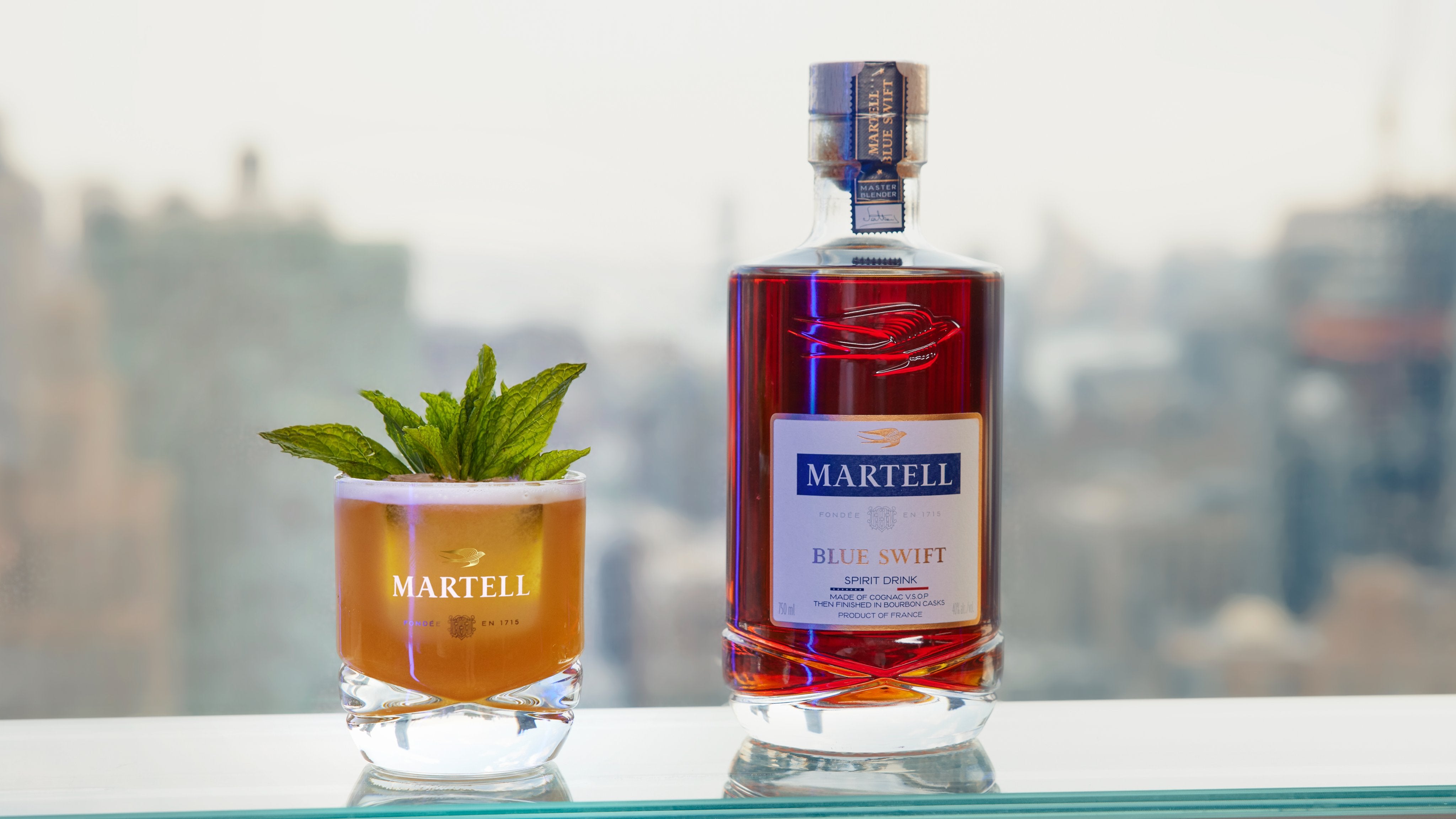 Martell Blue Swift Cognac bottle with a cocktail and mint garnish against a city skyline backdrop.