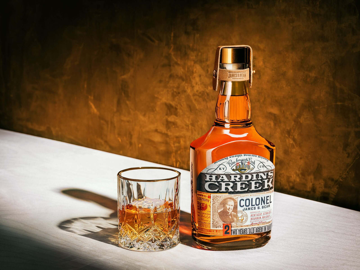 Hardin's Creek Colonel James B. Beam 2 Year Bourbon bottle next to a glass with bourbon on a white surface.
