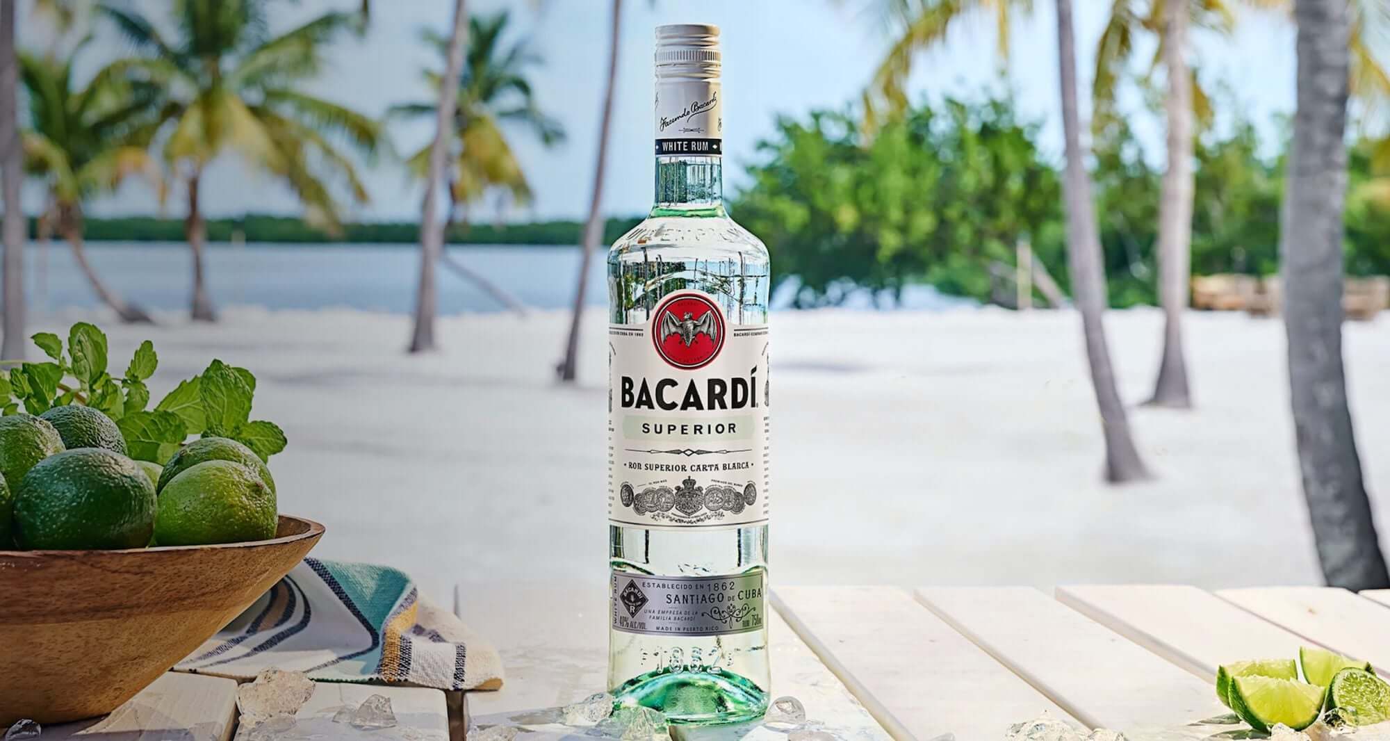 Bacardí Superior Rum bottle surrounded by limes on a beach table with palm trees in the background.