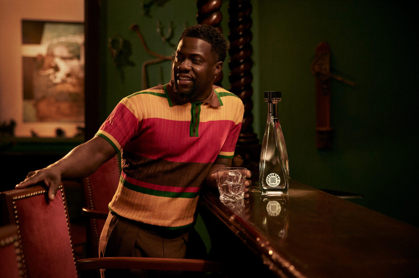 Kevin Hart enjoying a glass next to a bottle of Gran Coramino Cristalino Tequila in a vibrant bar setting.