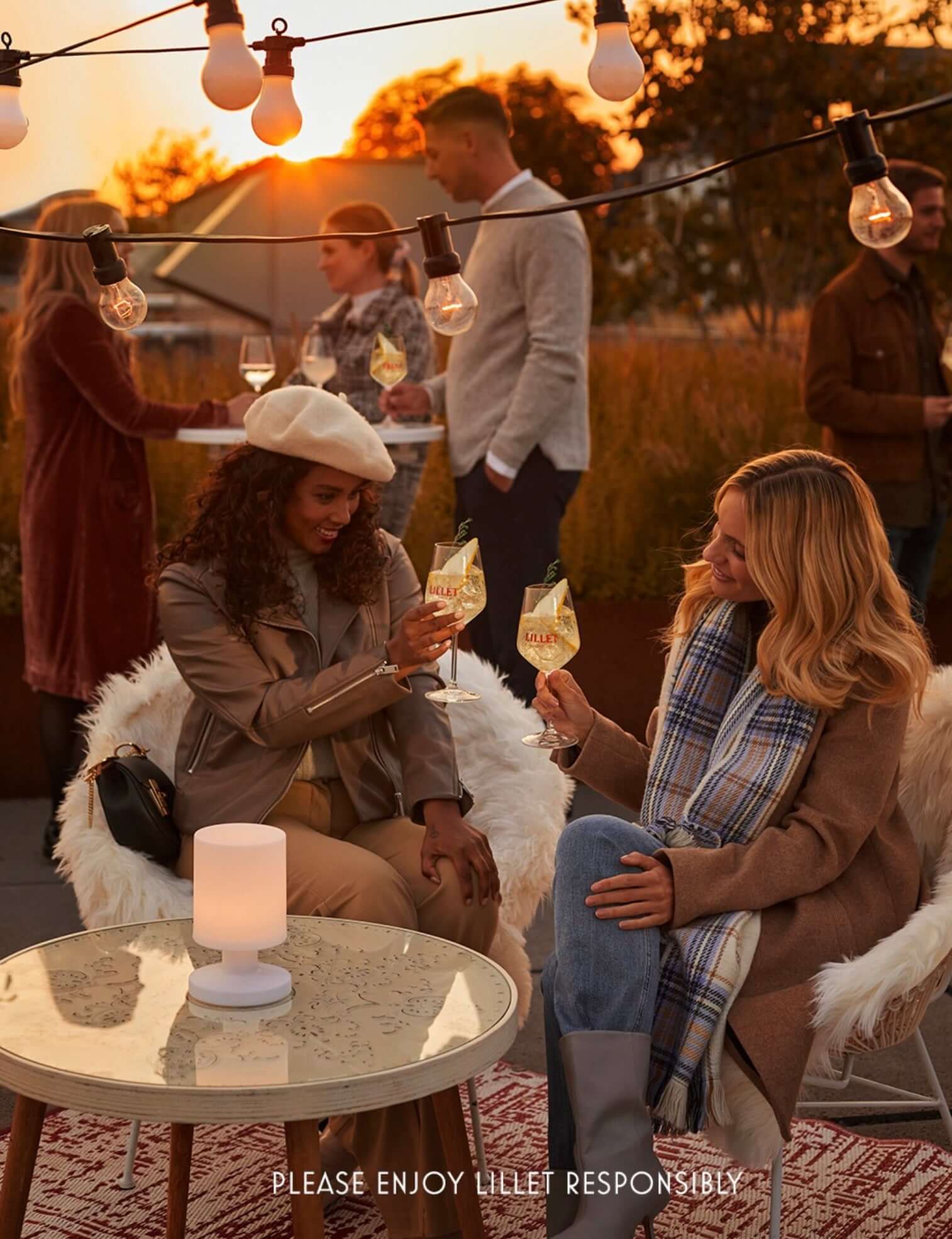 Two women clinking Lillet Aperitif Blanc glasses at sunset with friends in the background, promoting enjoyment and togetherness.