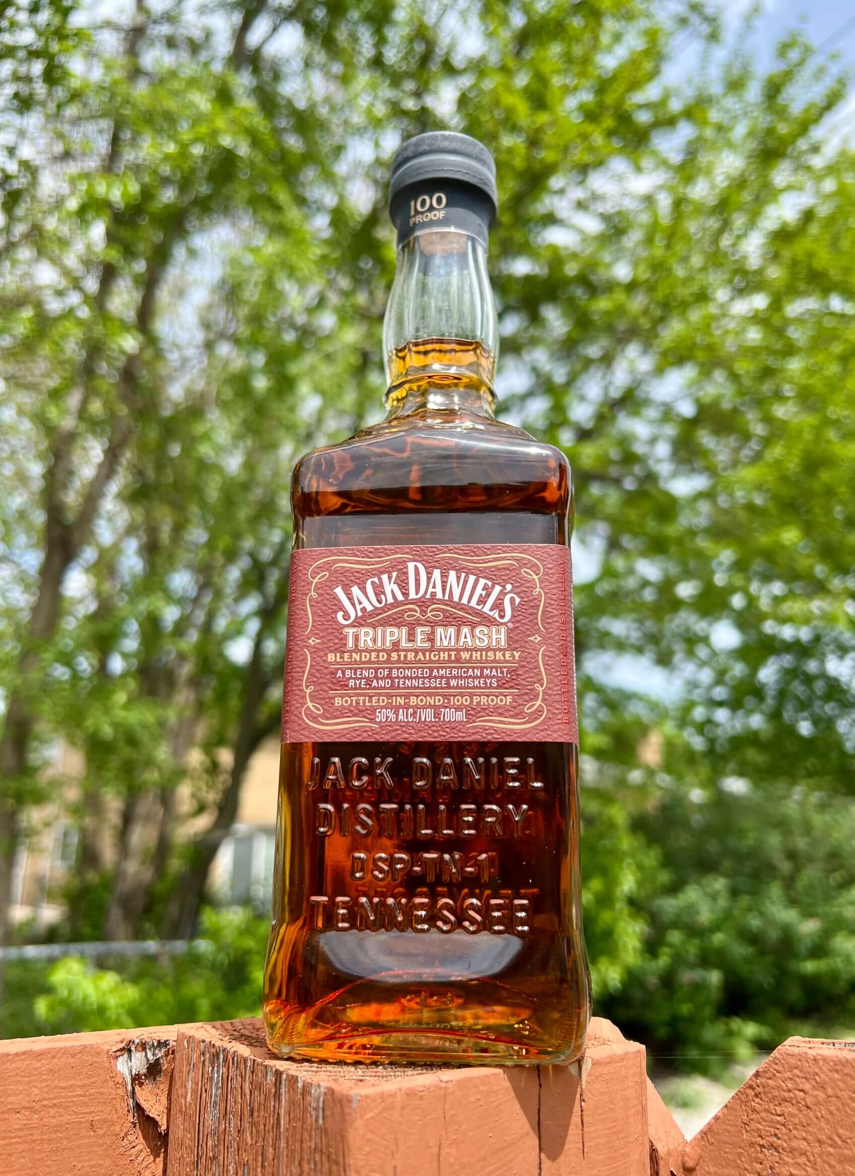 Jack Daniel's Triple Mash Bottled-In-Bond whiskey bottle with green trees in background, 100 proof, aged over four years.