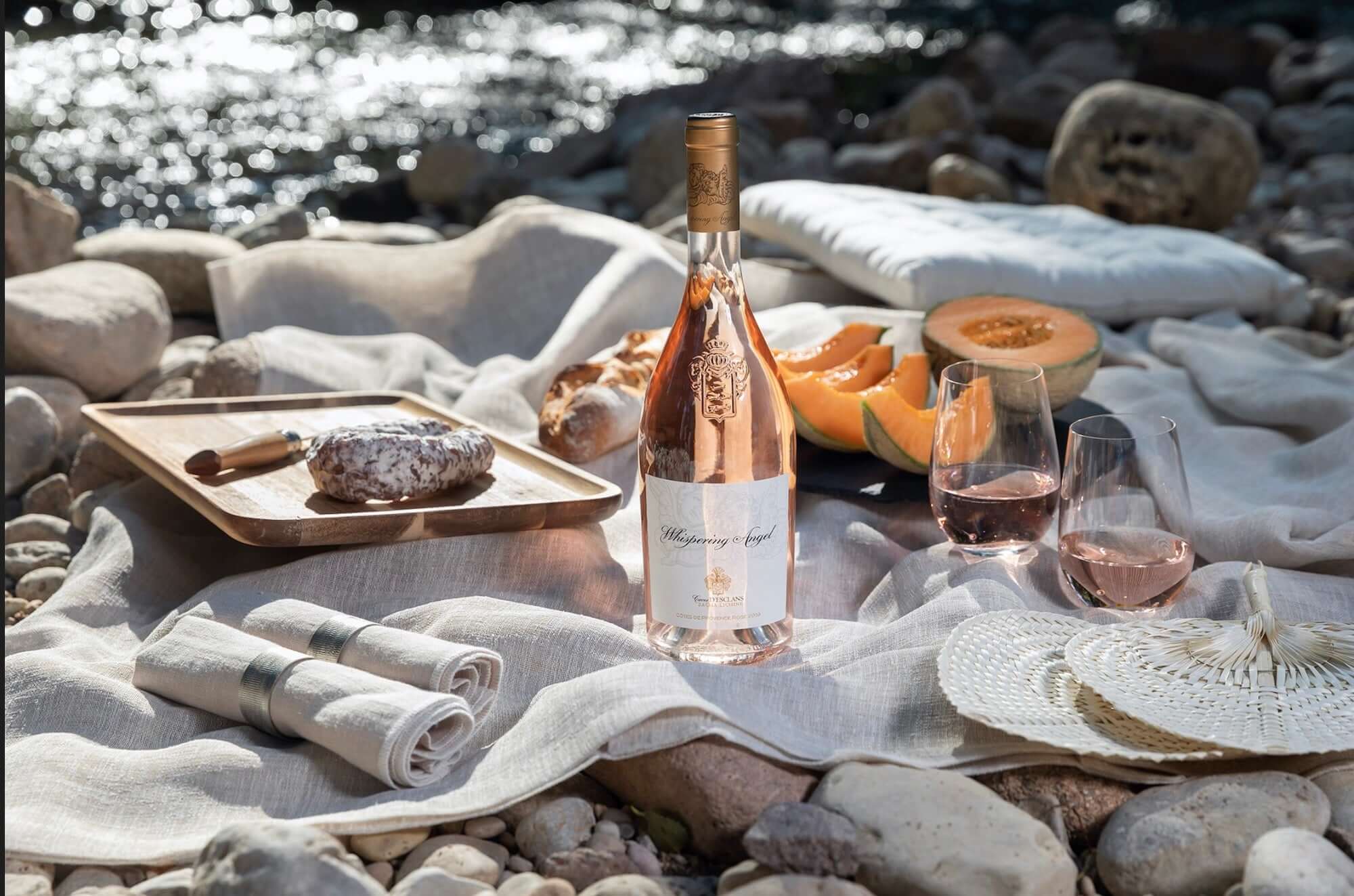 Château D'esclans Whispering Angel Rosé 750ml with picnic setting by the water featuring food and glasses of wine.