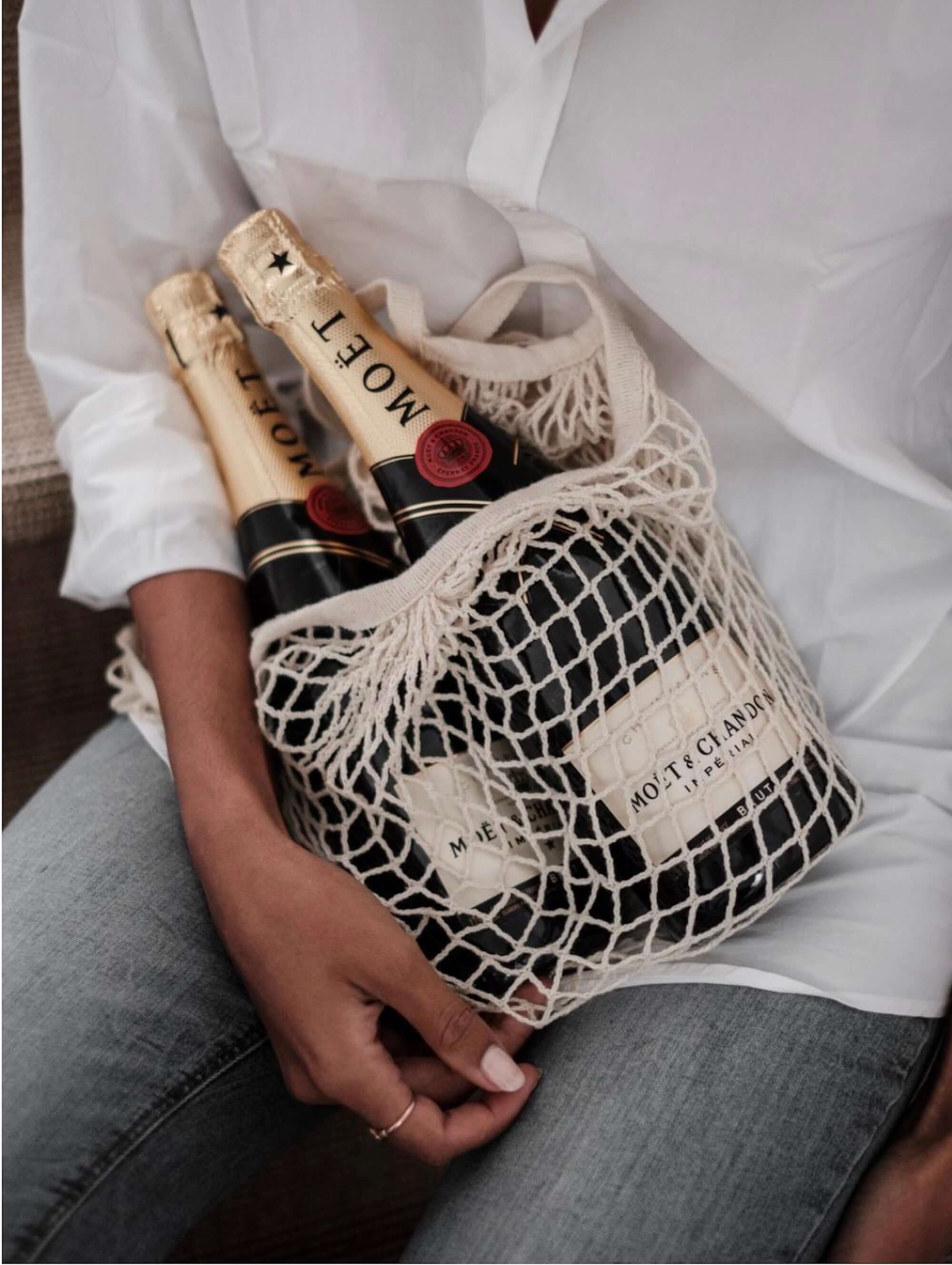 Moët & Chandon Brut Impérial champagne bottles in a reusable net tote bag, held by a person in casual attire.