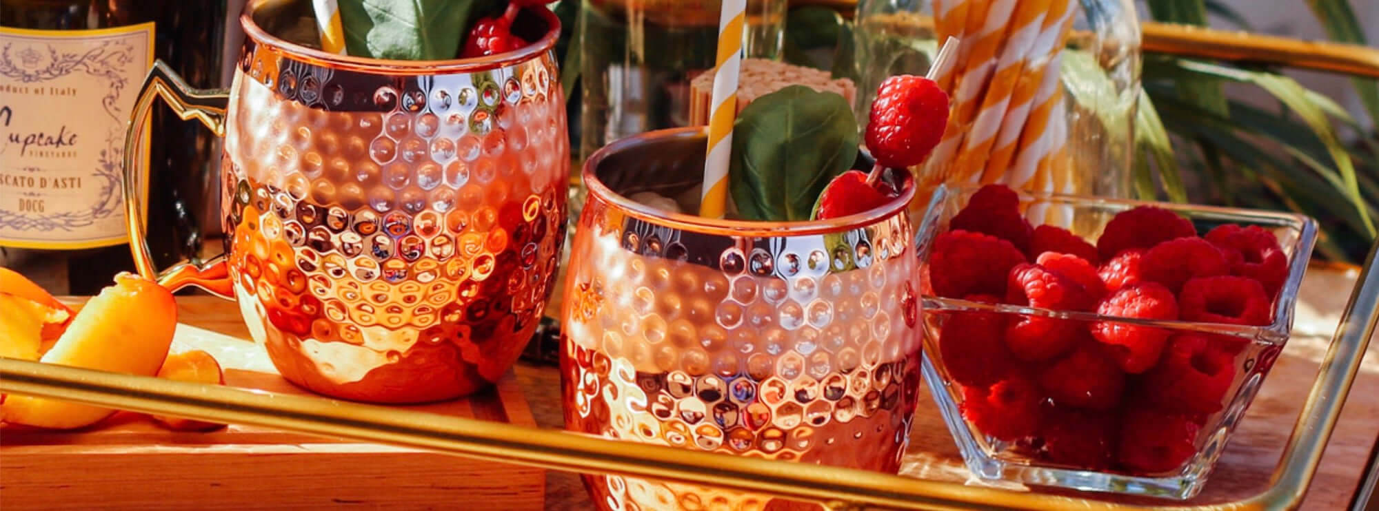 Stylish copper glasses filled with drinks, garnished with raspberries and mint, alongside fresh fruit on a serving tray.
