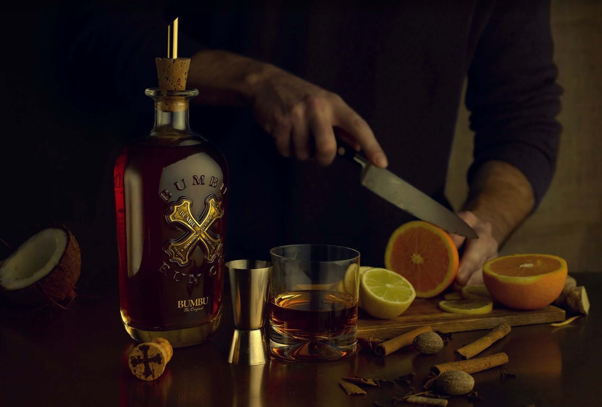 Bumbu The Original rum bottle beside a glass, fresh citrus, and spices on a wooden table.