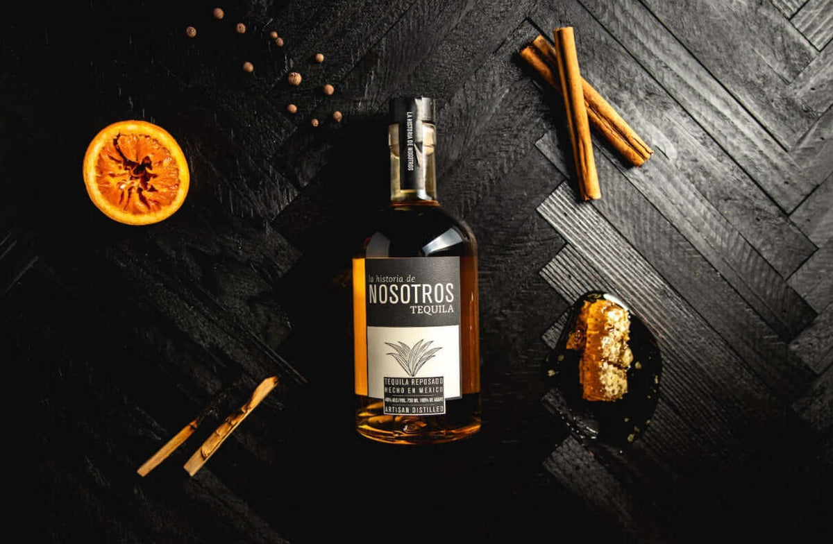 Bottle of Nosotros Reposado Tequila surrounded by orange, cinnamon sticks, and spices on a dark wooden surface.