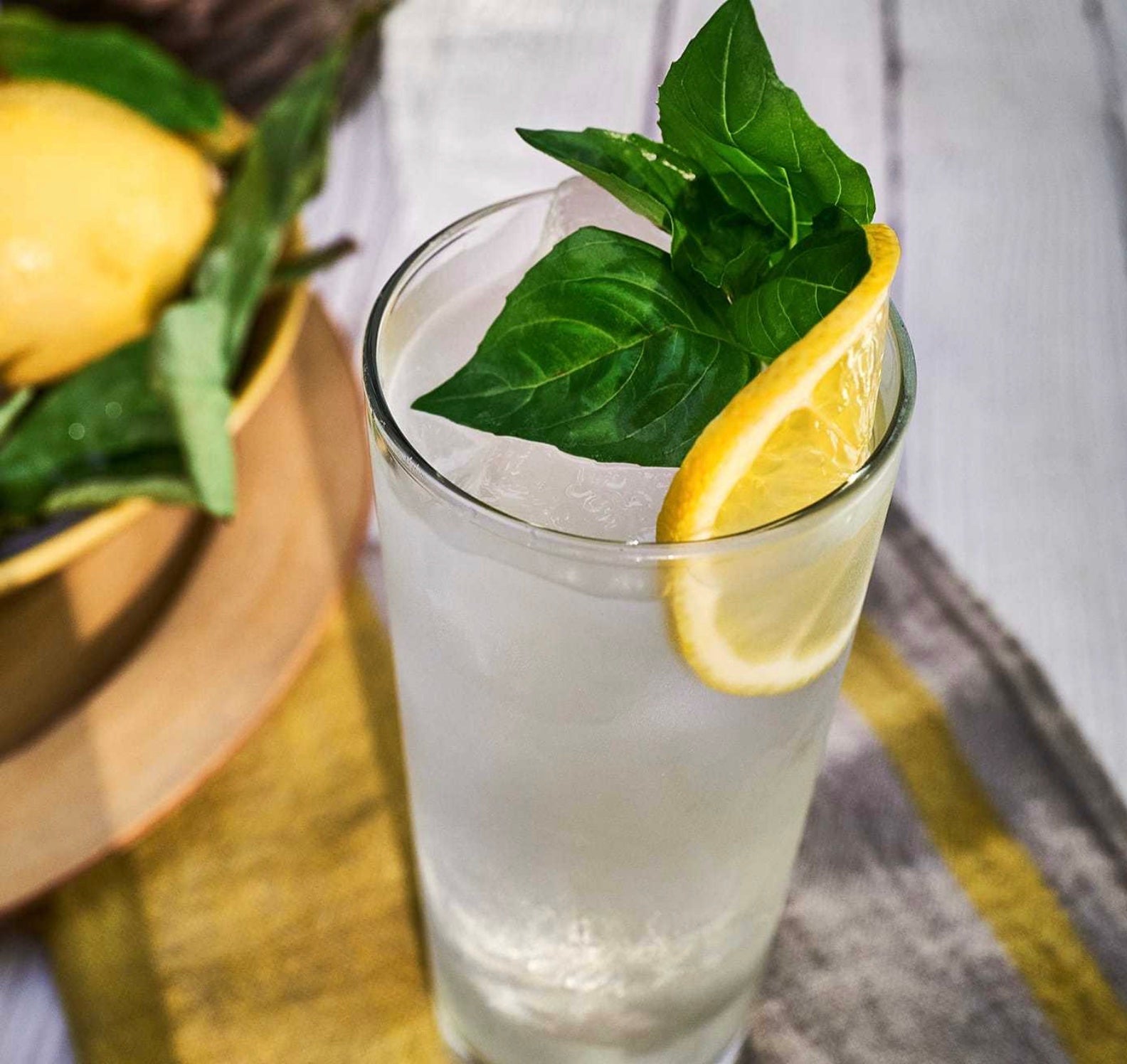 Refreshing cocktail with lemon and basil, served over ice in a tall glass, with fresh ingredients in the background.