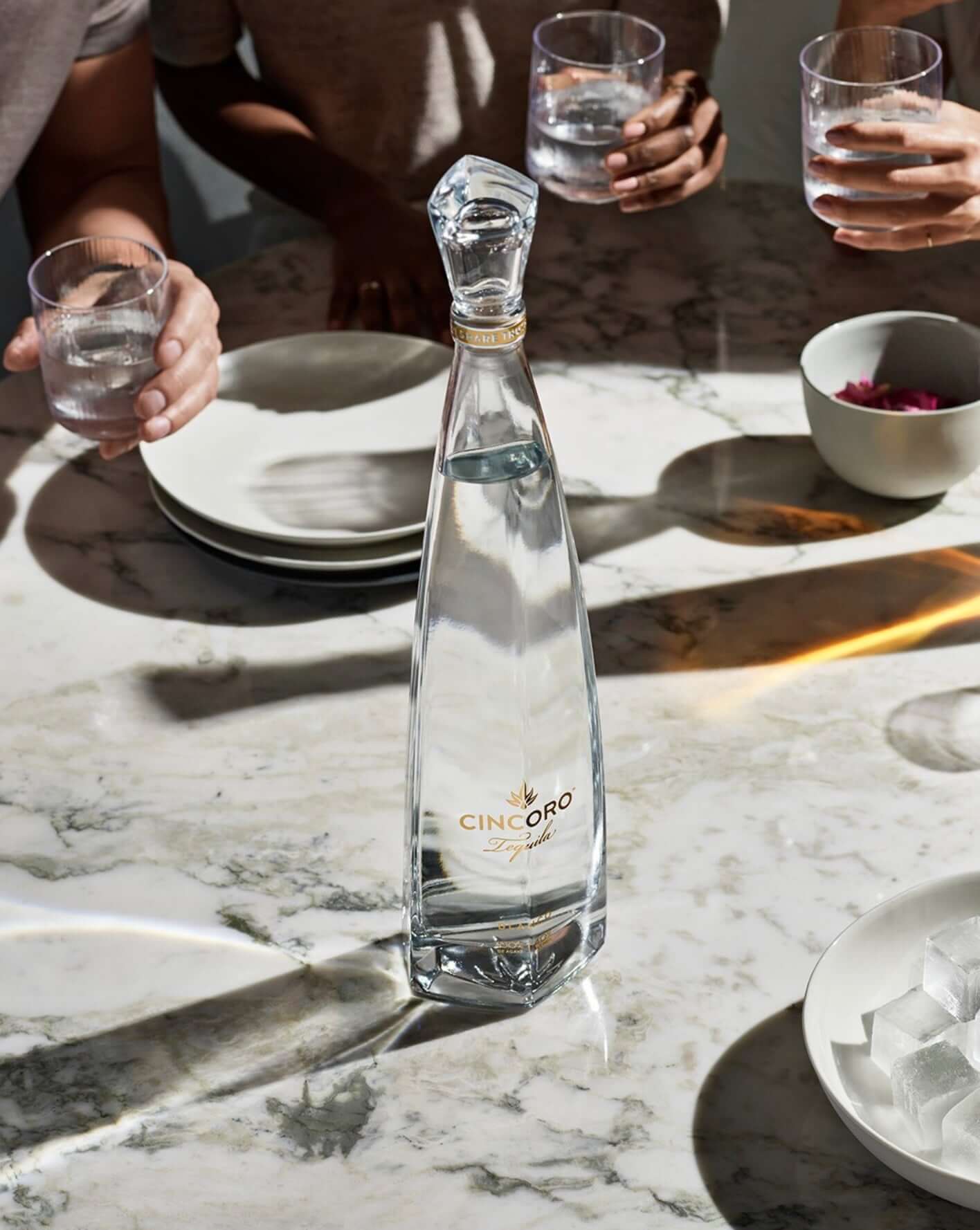 Cincoro Blanco Tequila bottle on a marble table surrounded by glasses and plates, showcasing its crystal-clear color.
