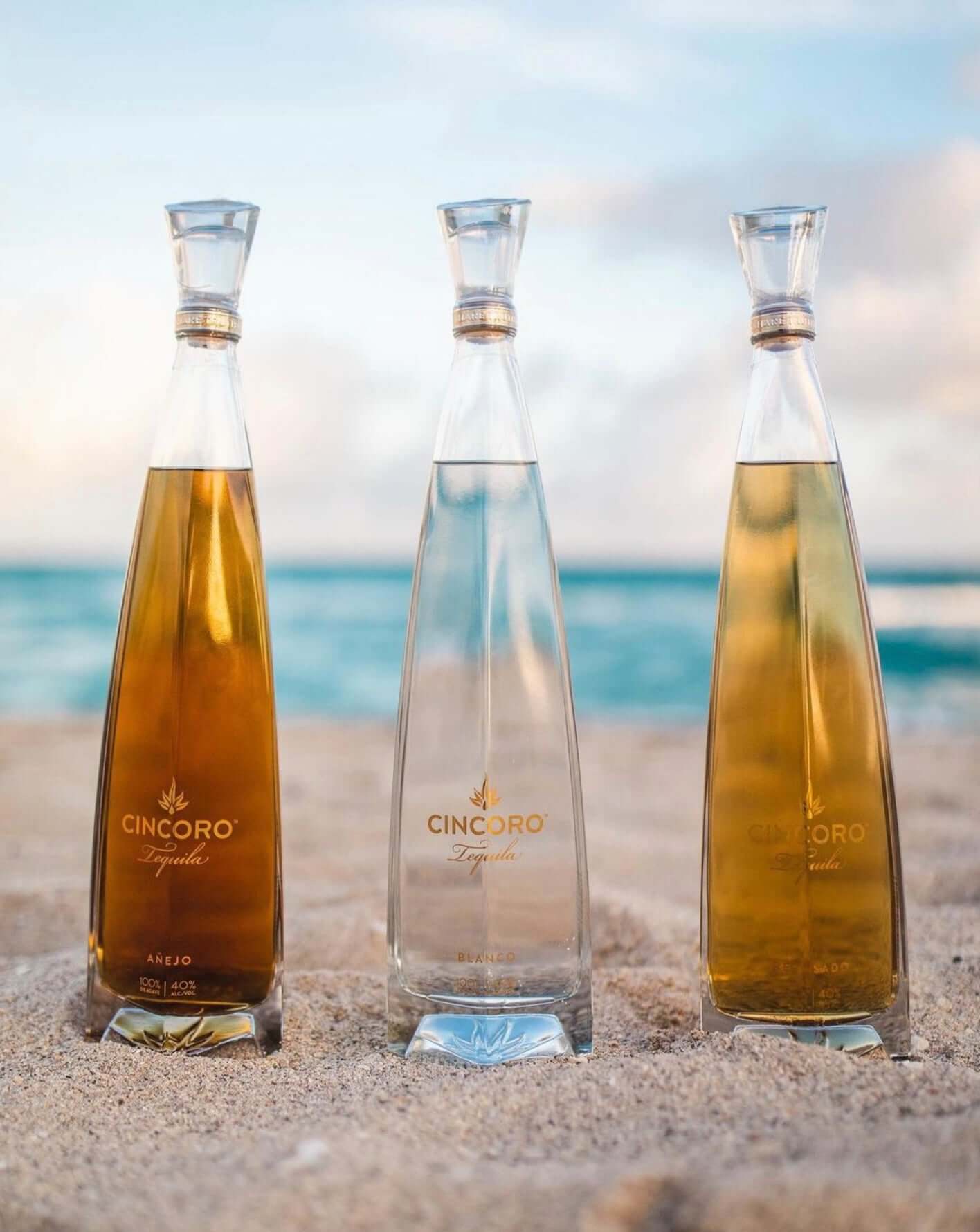 Cincoro Tequila bottles in different varieties arranged on sandy beach with ocean backdrop.