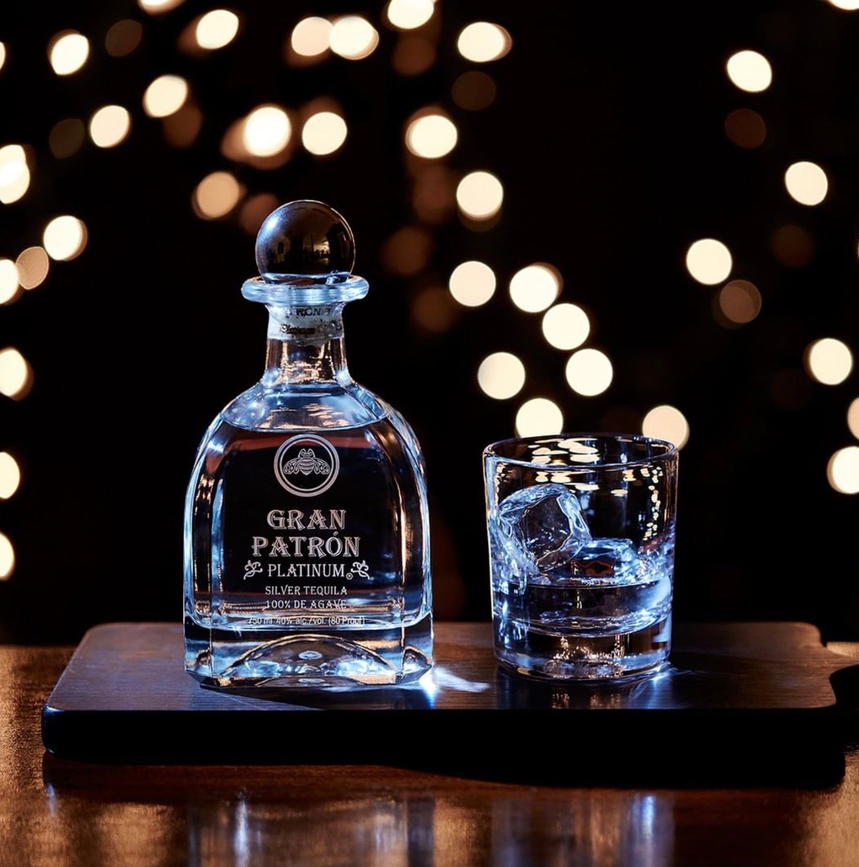 Gran Patrón Platinum Tequila in a crystal bottle with a glass on a wooden tray, sparkling bokeh background.