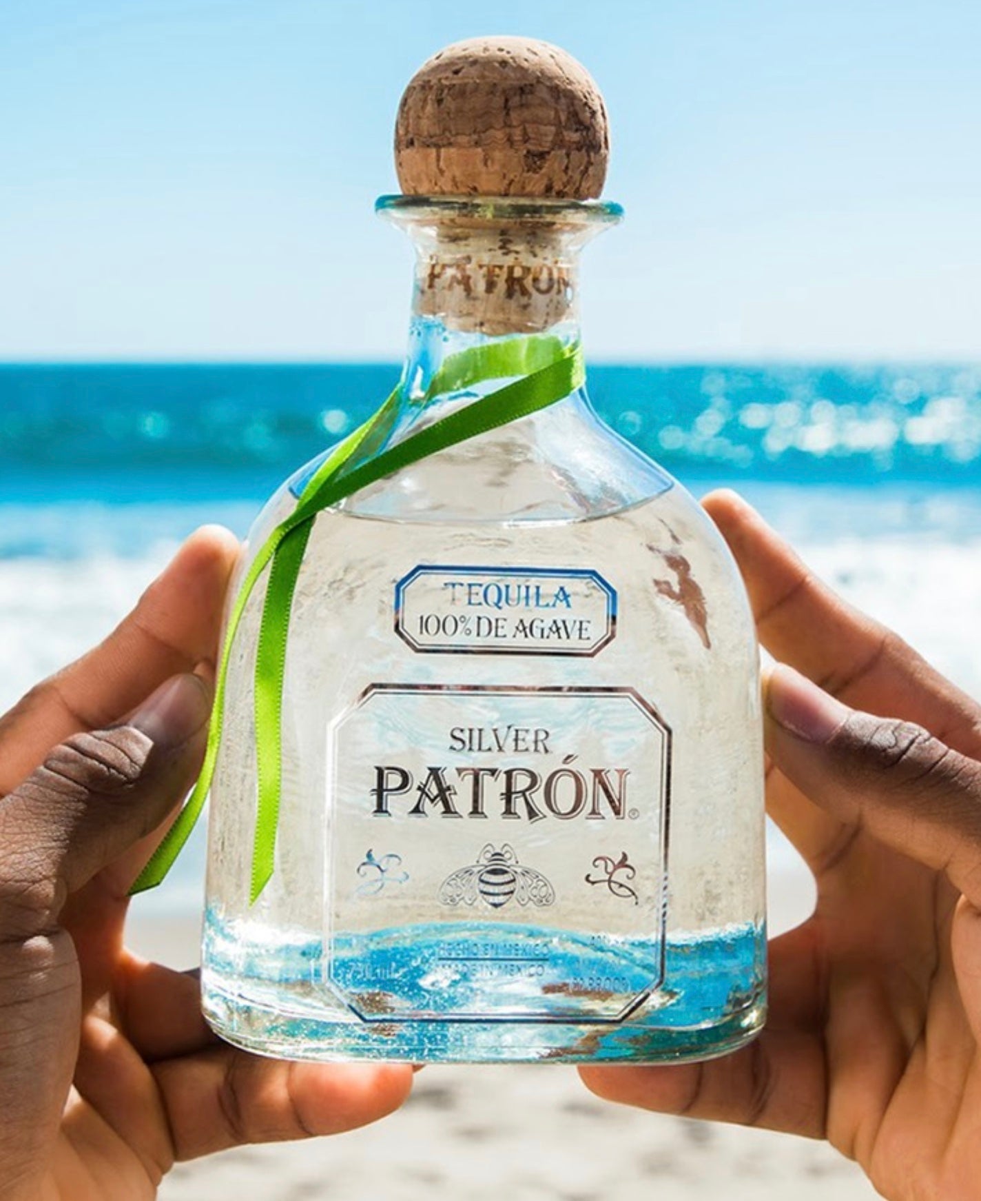 A hand holding a bottle of Patrón Silver Tequila at the beach, showcasing its crystal clear appearance and vibrant label.