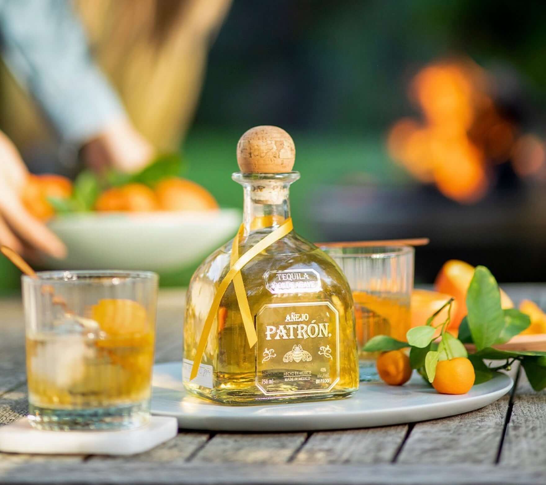 Patrón Añejo Tequila bottle on a table with cocktails and fresh fruit, highlighting its rich amber color and premium packaging.