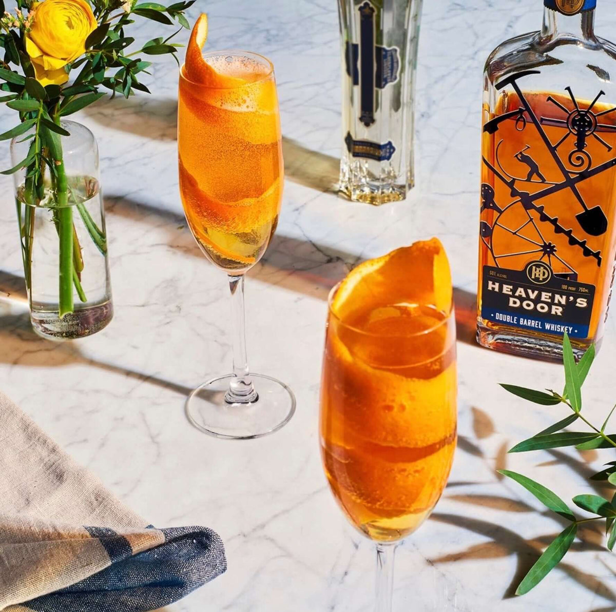 Two elegant cocktails with orange twists beside Heaven's Door Double Barrel Whiskey bottle on a marble table.