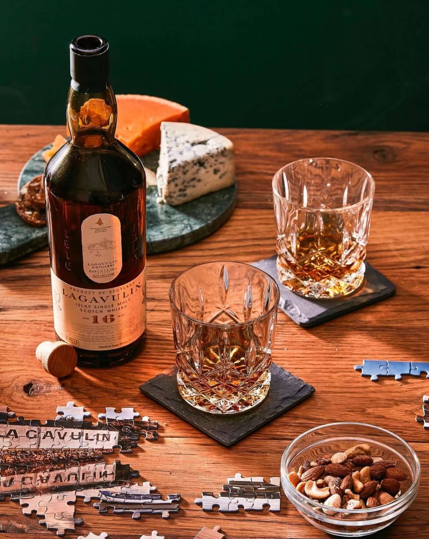 Lagavulin 16 Year whiskey bottle with glasses, cheese, nuts, and puzzle pieces on a wooden table.