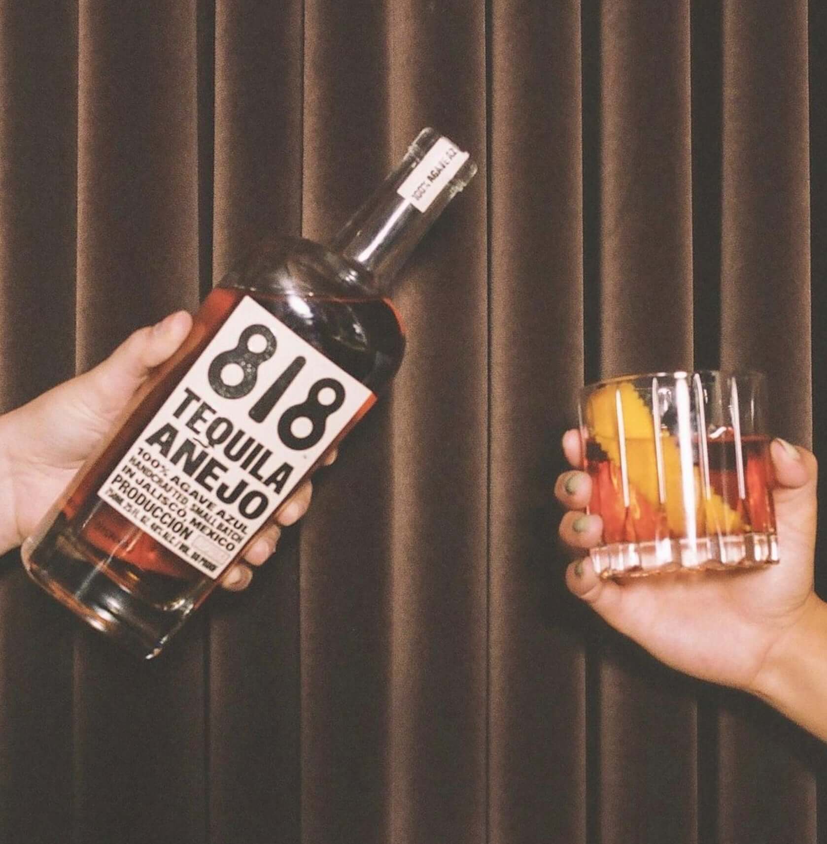 A hand holding a bottle of 818 Añejo Tequila next to a glass of mixed fruit cocktail.