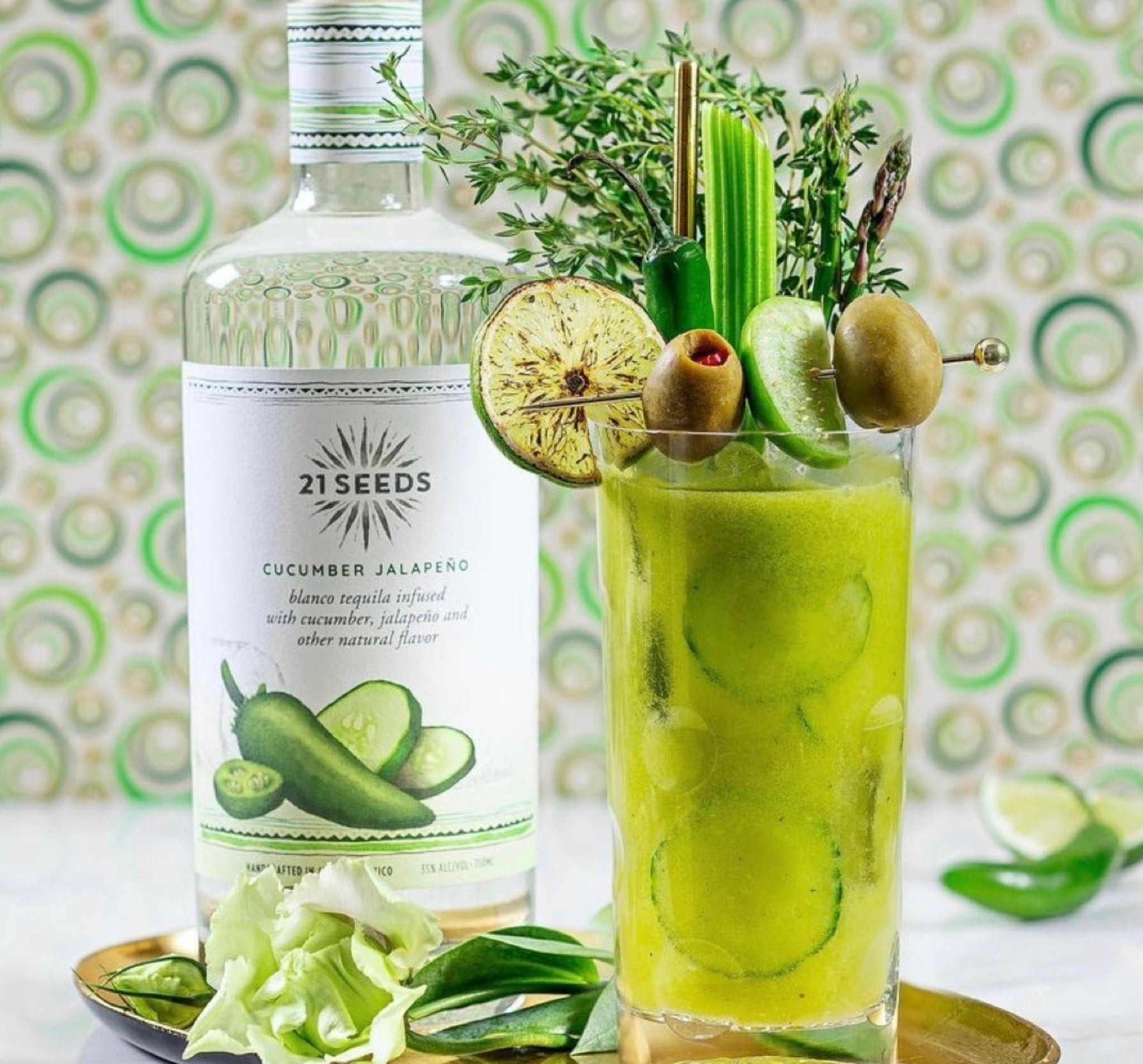Glass of Cucumber Jalapeño cocktail next to 21 Seeds tequila bottle, garnished with herbs and fresh ingredients.
