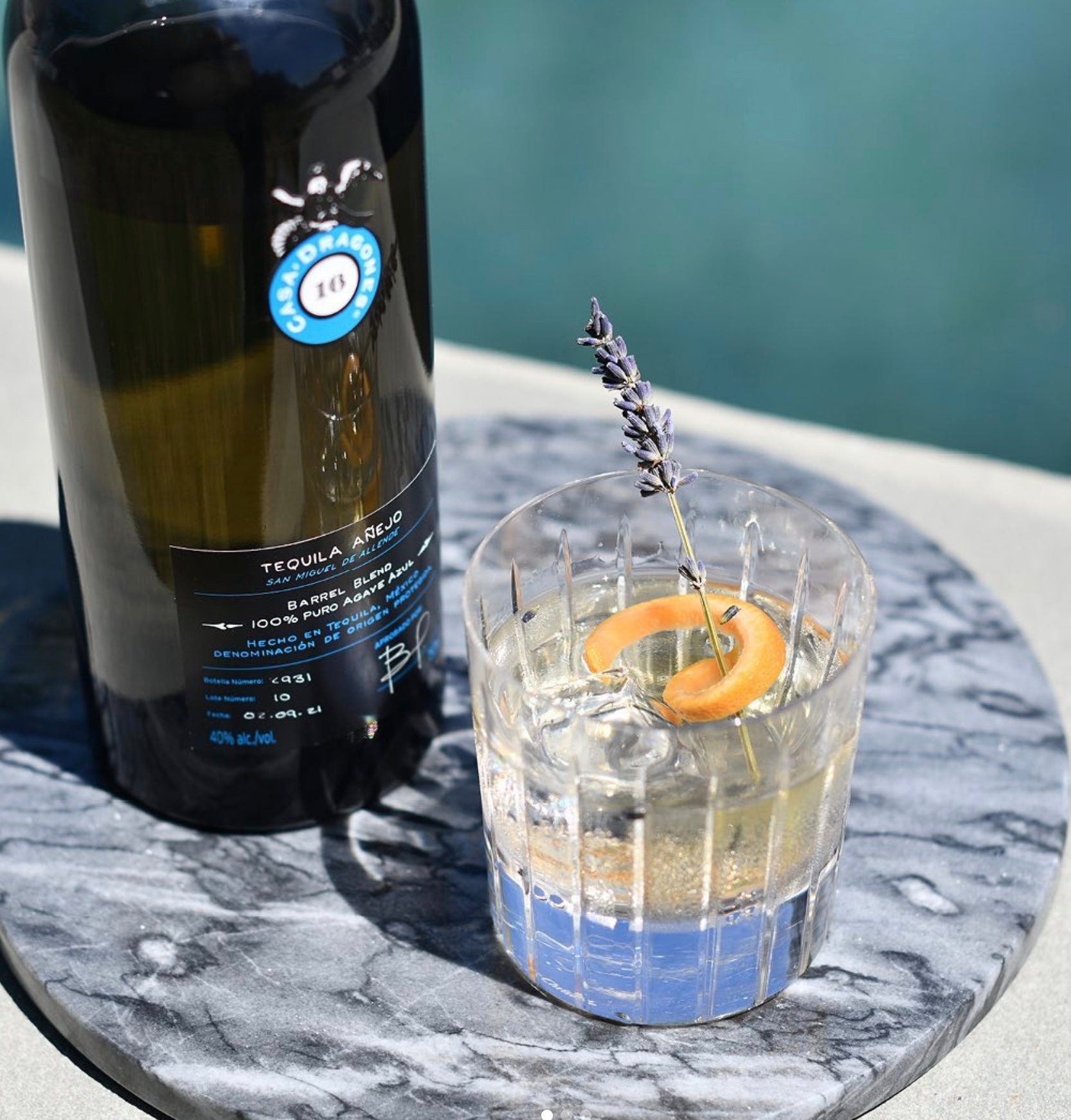 Casa Dragones Añejo Barrel Blend Tequila bottle beside a cocktail garnished with lavender and orange slice on a marble surface.