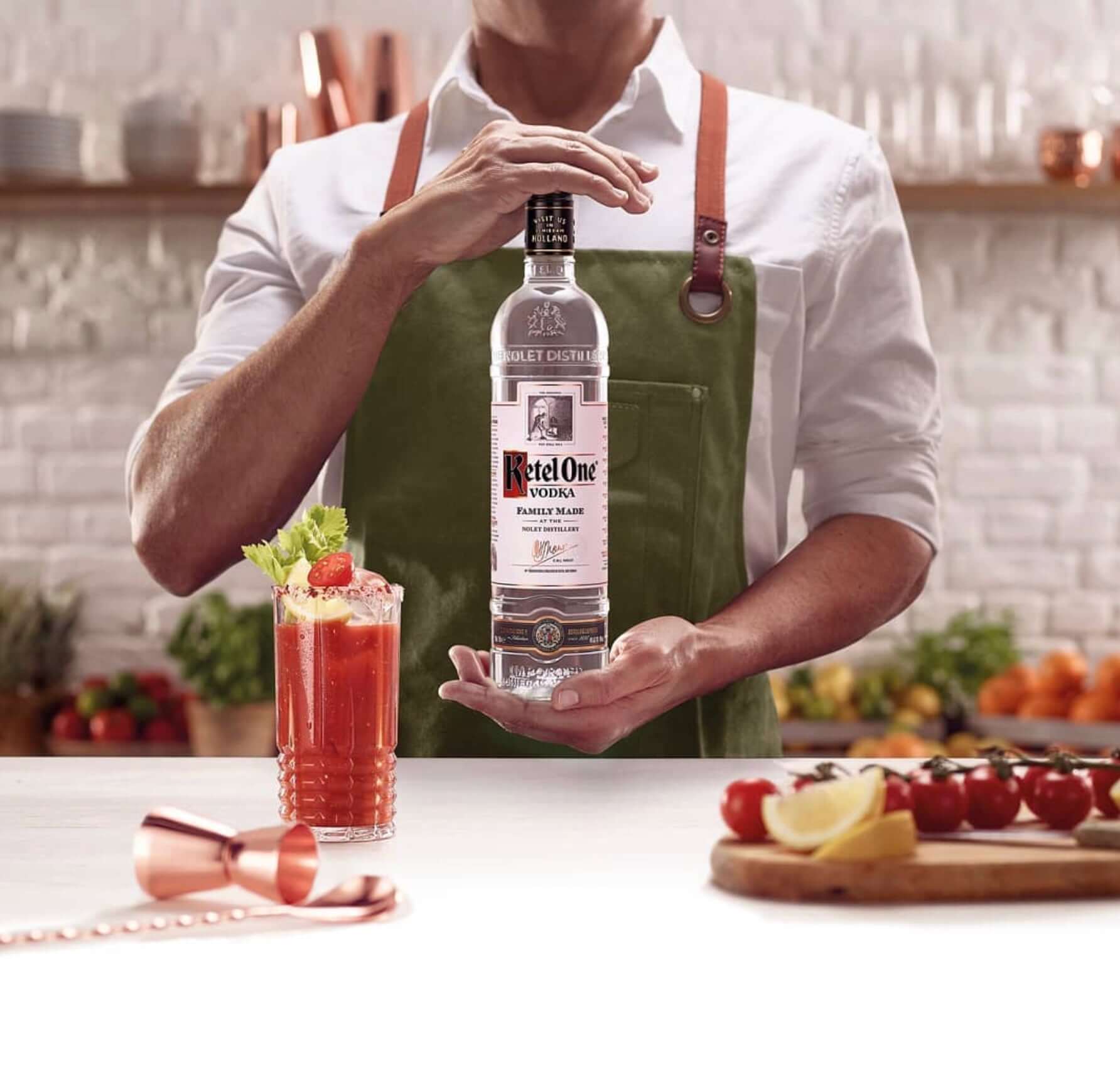 Person in apron holding a bottle of Ketel One Vodka beside a Bloody Mary cocktail on a kitchen counter.