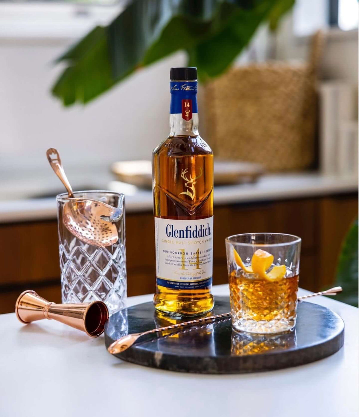 Glenfiddich 14 Year Bourbon Barrel Reserve bottle with whiskey glass, cocktail tools on a stylish kitchen countertop.