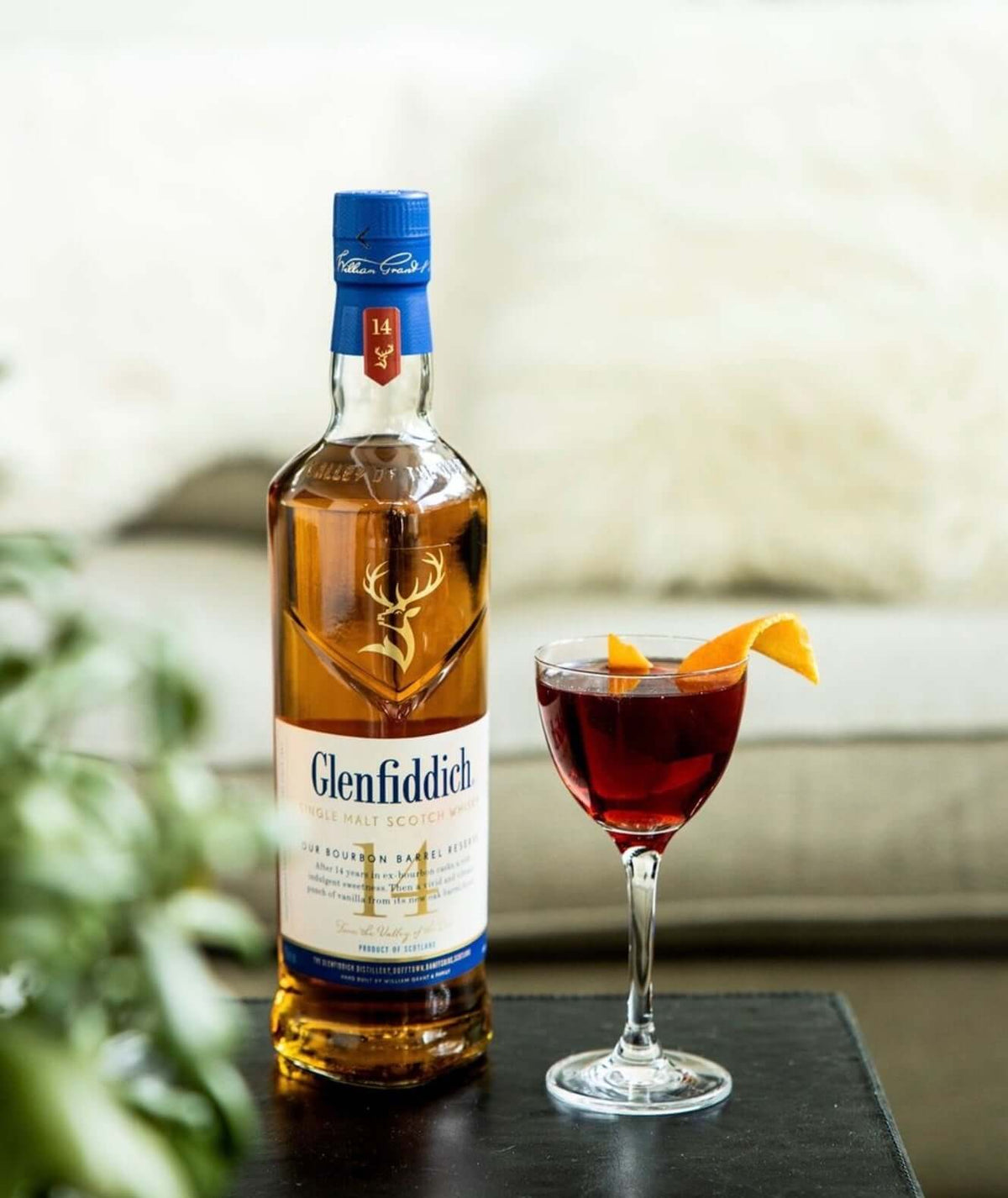 Glenfiddich 14 Year Bourbon Barrel Reserve bottle next to a cocktail with orange garnish on a table.