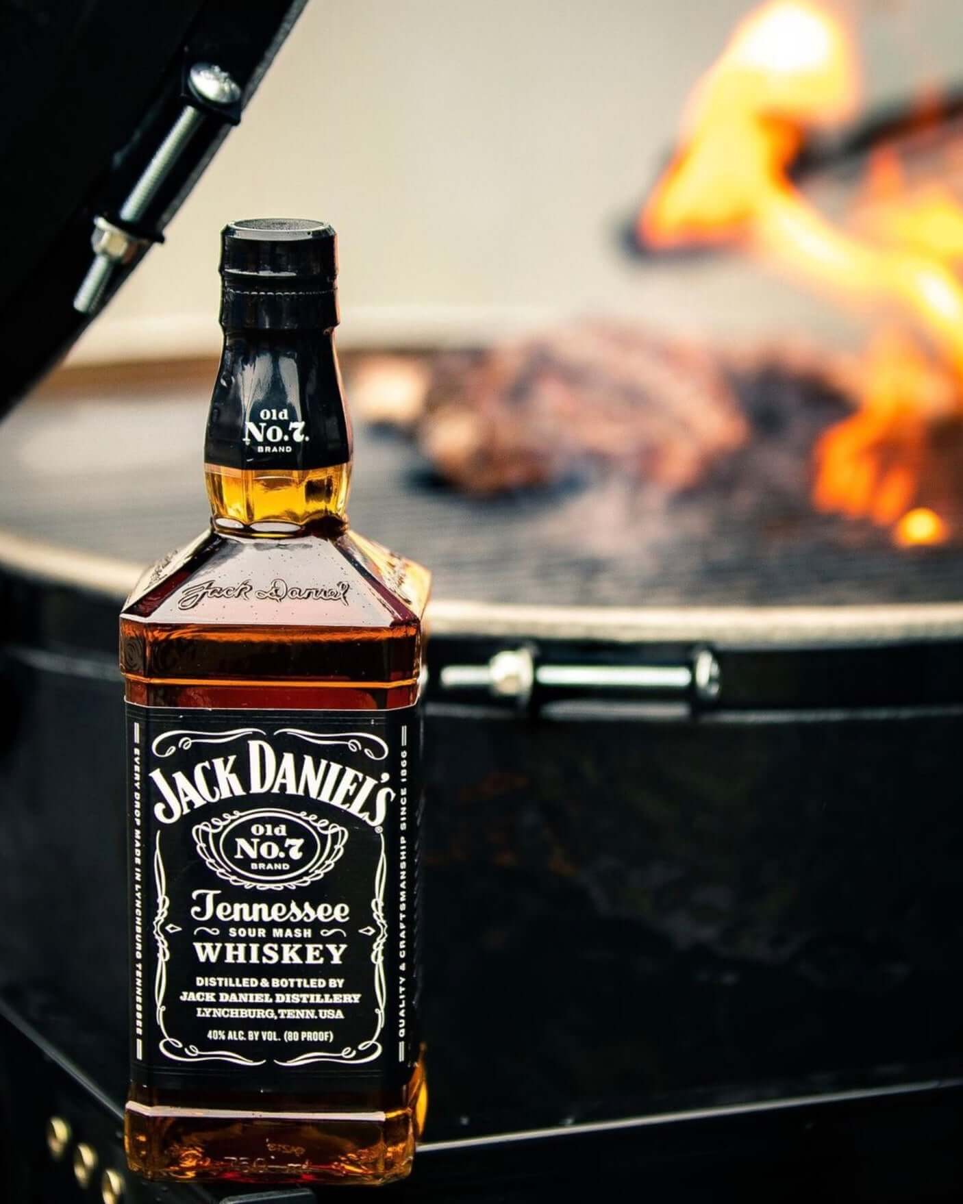 Jack Daniel's Tennessee Whiskey bottle by a grill with fire, highlighting its charcoal mellowing and handcrafted quality.