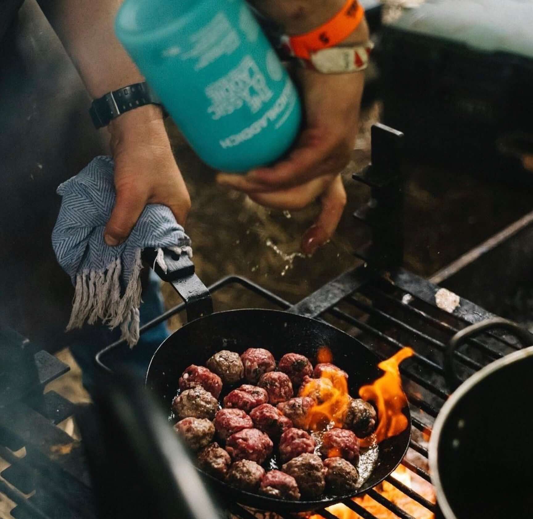 Grilling meatballs over an open flame in a cast iron pan, with a hand sprinkling seasoning.