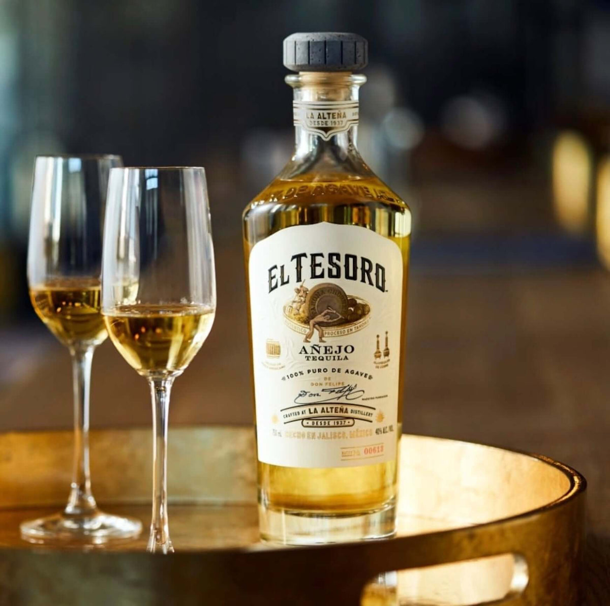 El Tesoro Añejo Tequila bottle with two elegant glasses on a golden tray, showcasing its rich gold color.