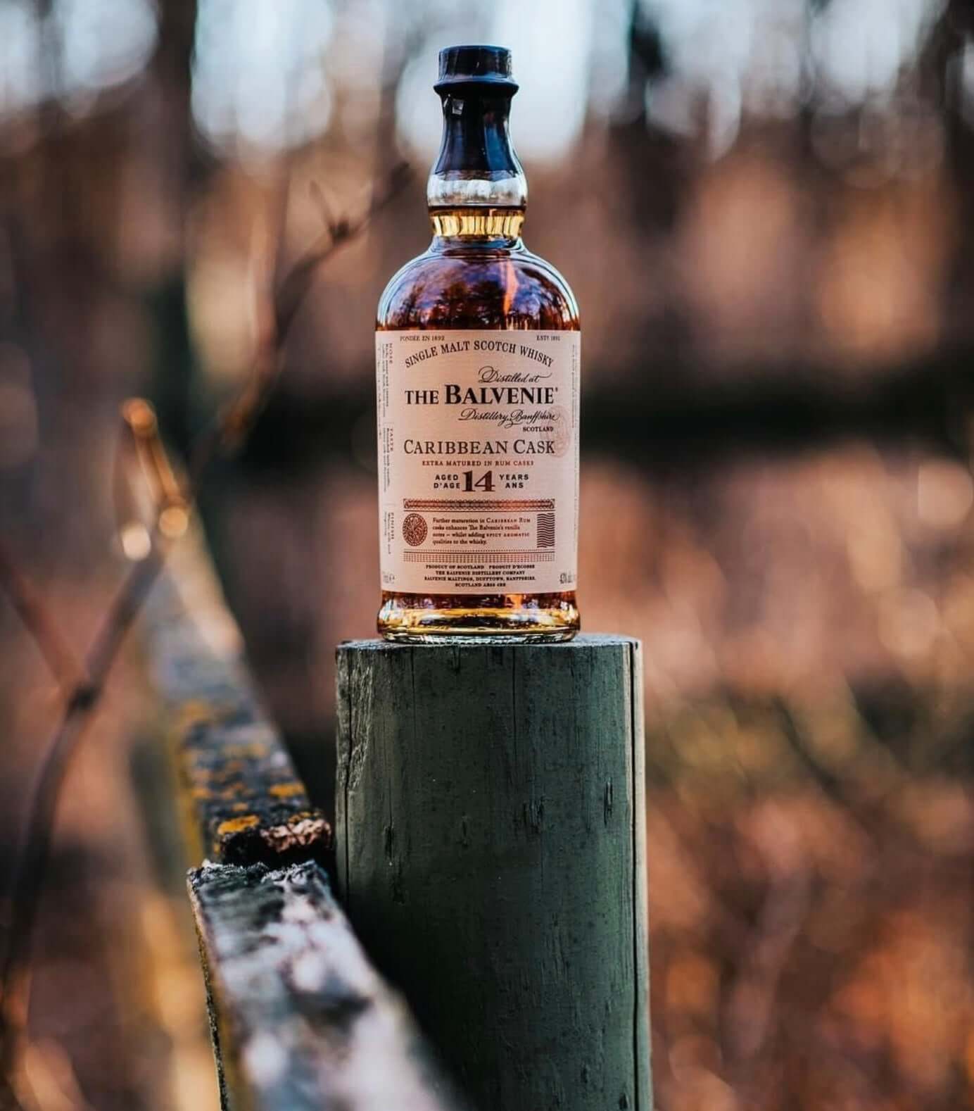 The Balvenie Caribbean Cask 14 Year single malt whisky bottle on a wooden post in a natural setting.