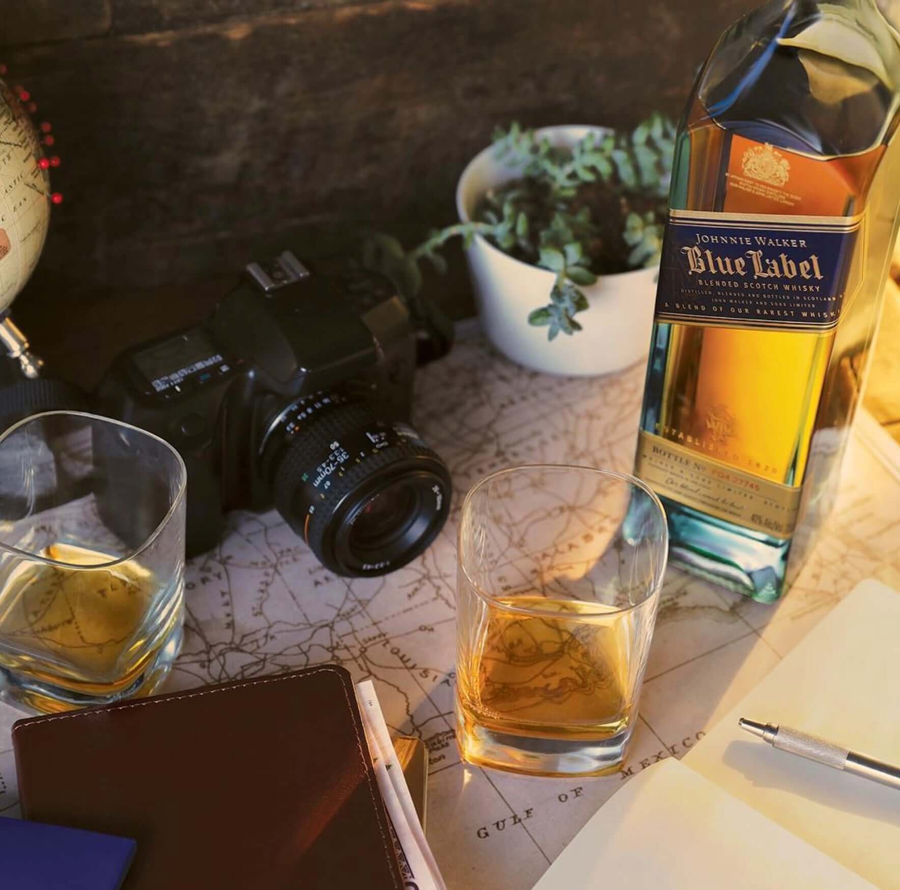 Photo of Johnnie Walker Blue Label whisky bottle with glasses, camera, and globe on a vintage map.