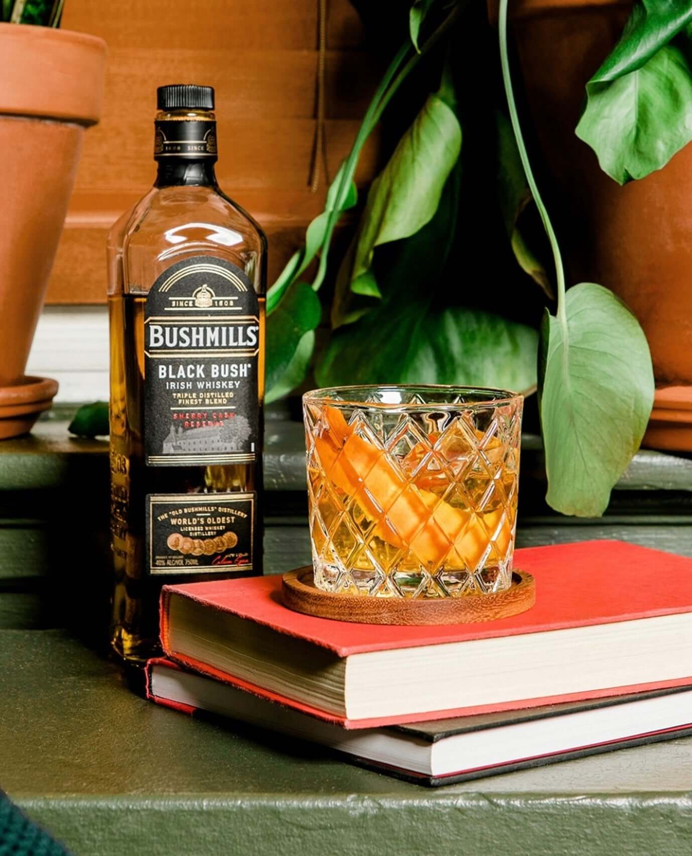 Bushmills Black Bush Irish Whiskey bottle with a cocktail featuring an orange slice beside books and greenery.