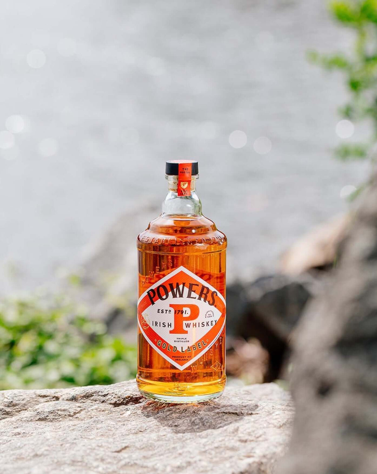 Powers Gold Label Irish Whiskey bottle on a rock by the water, showcasing its bold amber color and distinctive label.
