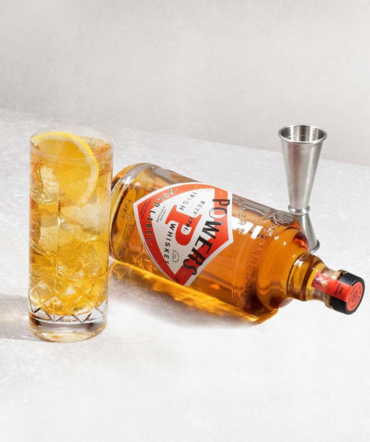 Powers Gold Label Irish Whiskey bottle with a cocktail and a jigger, showcasing its bold character and vibrant presentation.