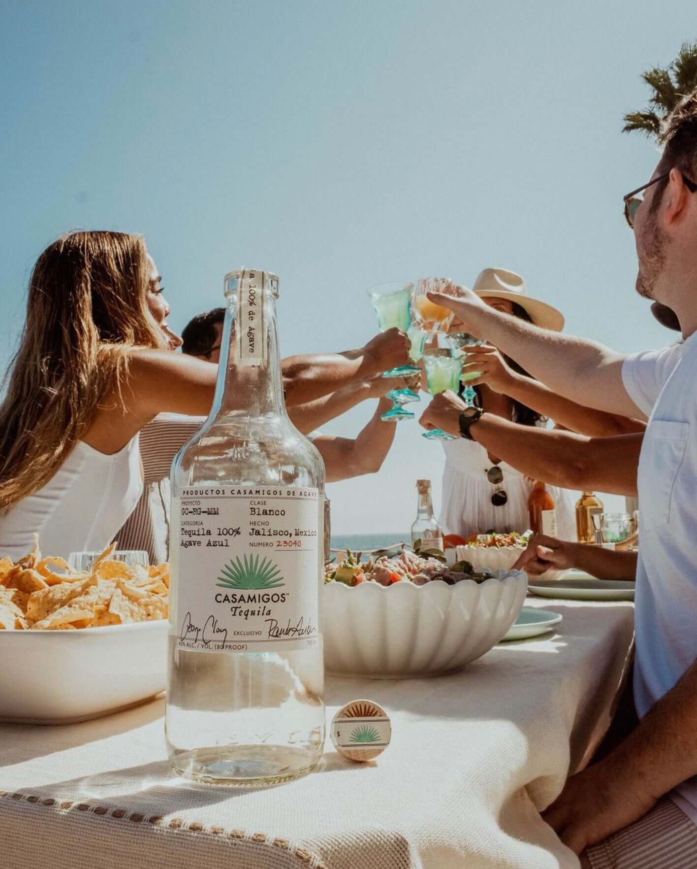 Friends toasting with Casamigos Blanco Tequila at a sunny outdoor gathering with snacks.