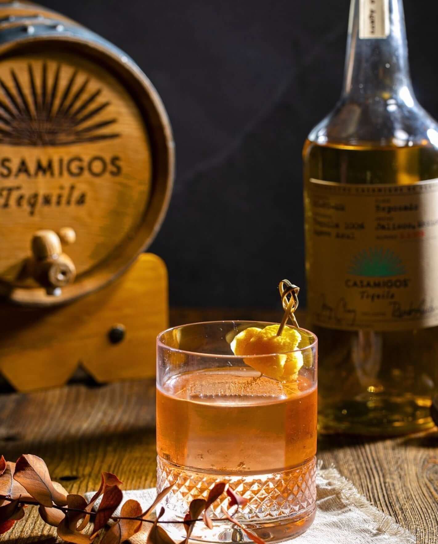 A glass of Casamigos Reposado Tequila cocktail with garnish, featuring a bottle and barrel in the background.