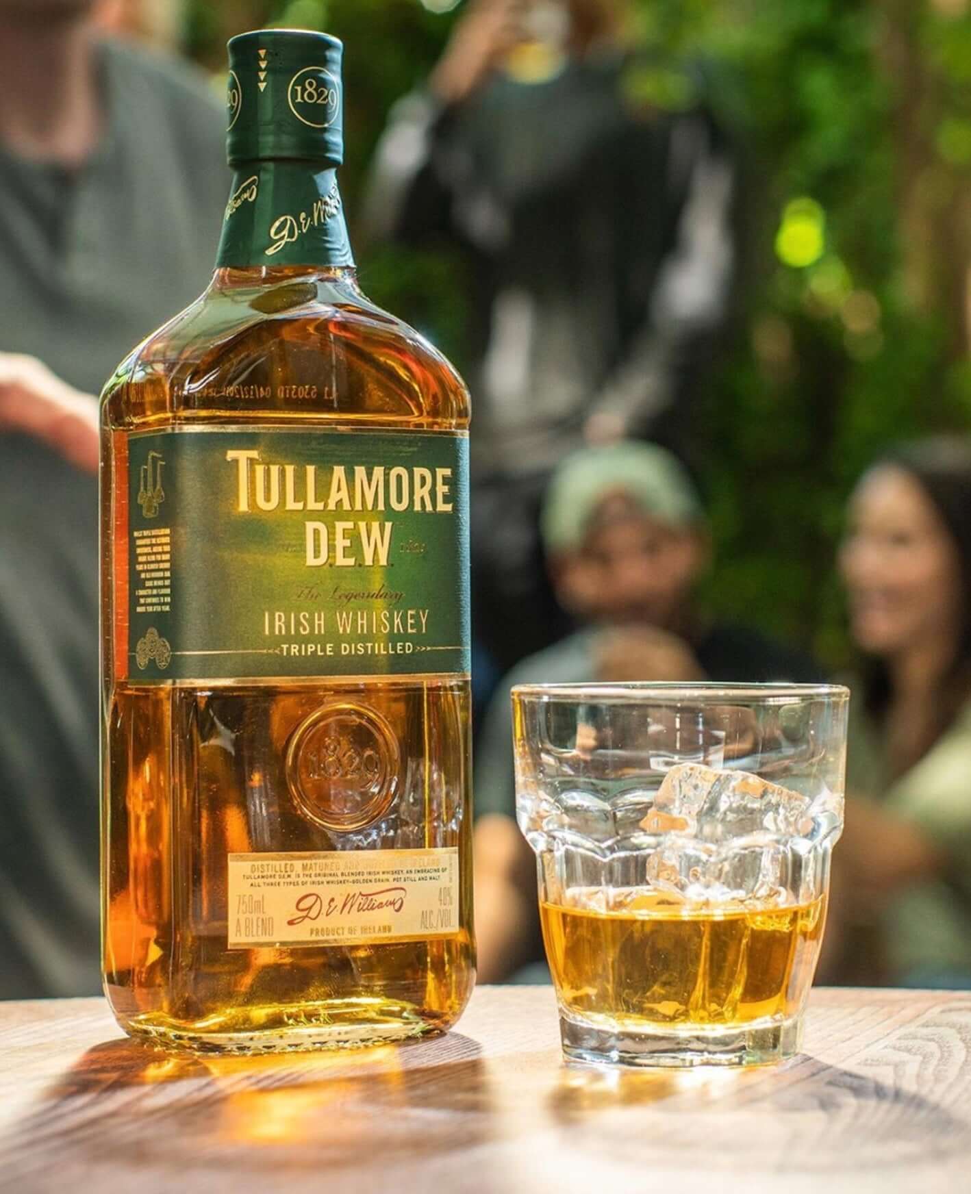 Tullamore Dew Original Irish Whiskey bottle with a glass of whiskey on ice, set in a lively outdoor gathering.