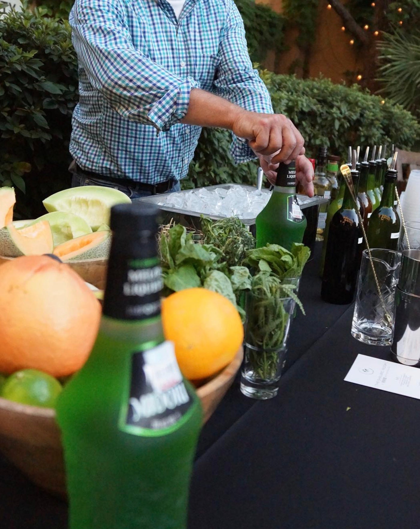 Bartender preparing cocktails with Midori Melon Liqueur and fresh fruits at an outdoor event.