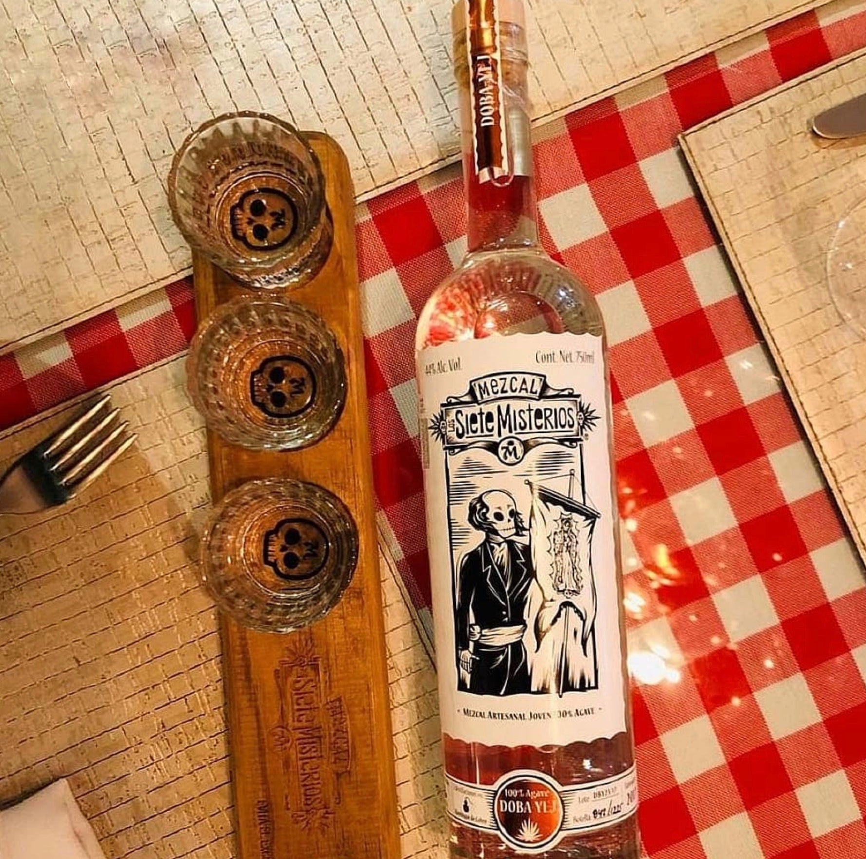 Siete Misterios Mezcal Doba-Yej bottle with tasting glasses on a checkered tablecloth.