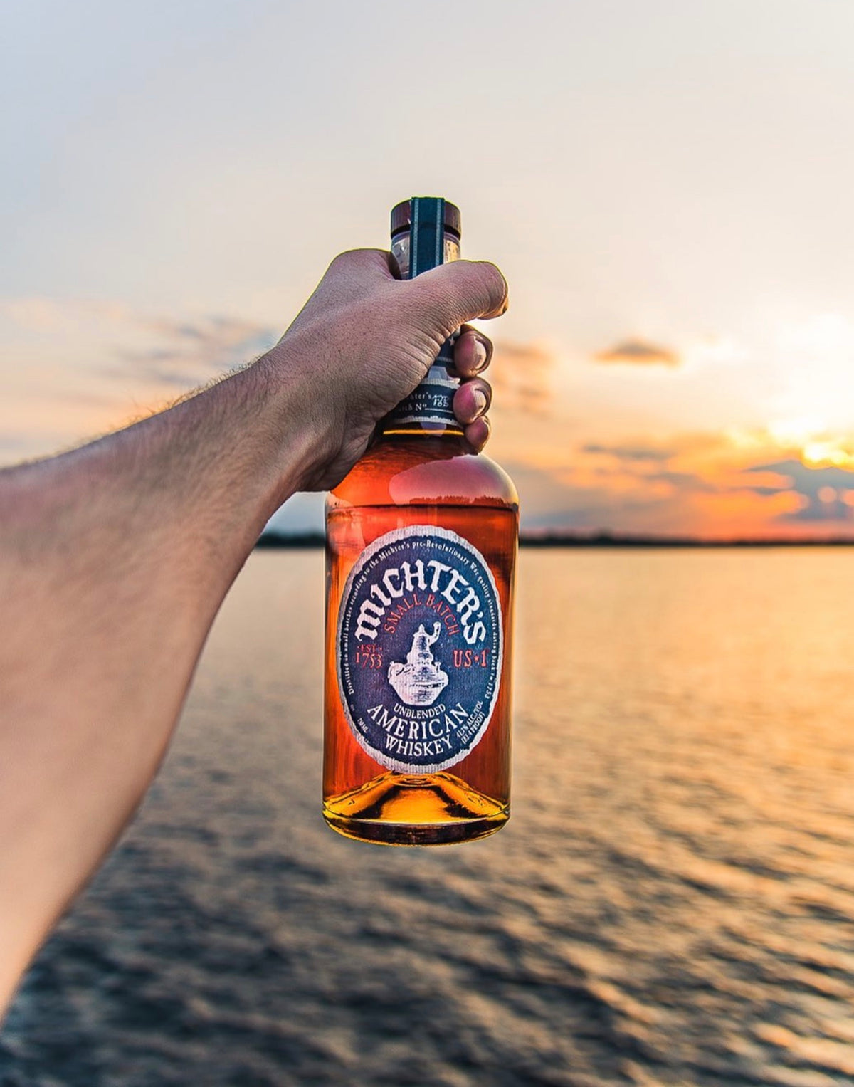 A hand holding a bottle of Michter's Unblended American Whiskey at sunset by the water.