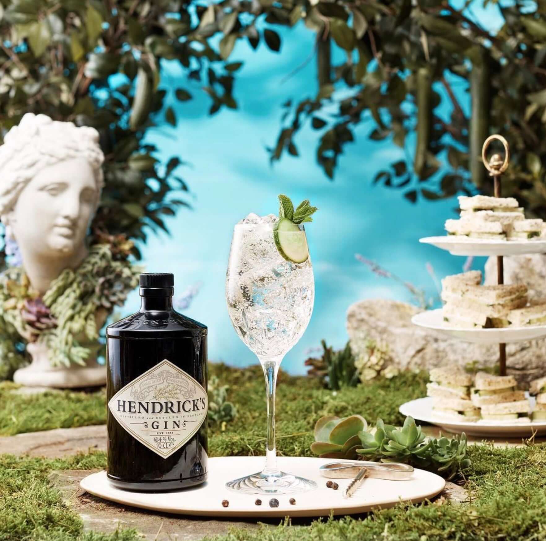 Hendrick's Gin bottle with a refreshing cocktail garnished with cucumber and mint, surrounded by nature.