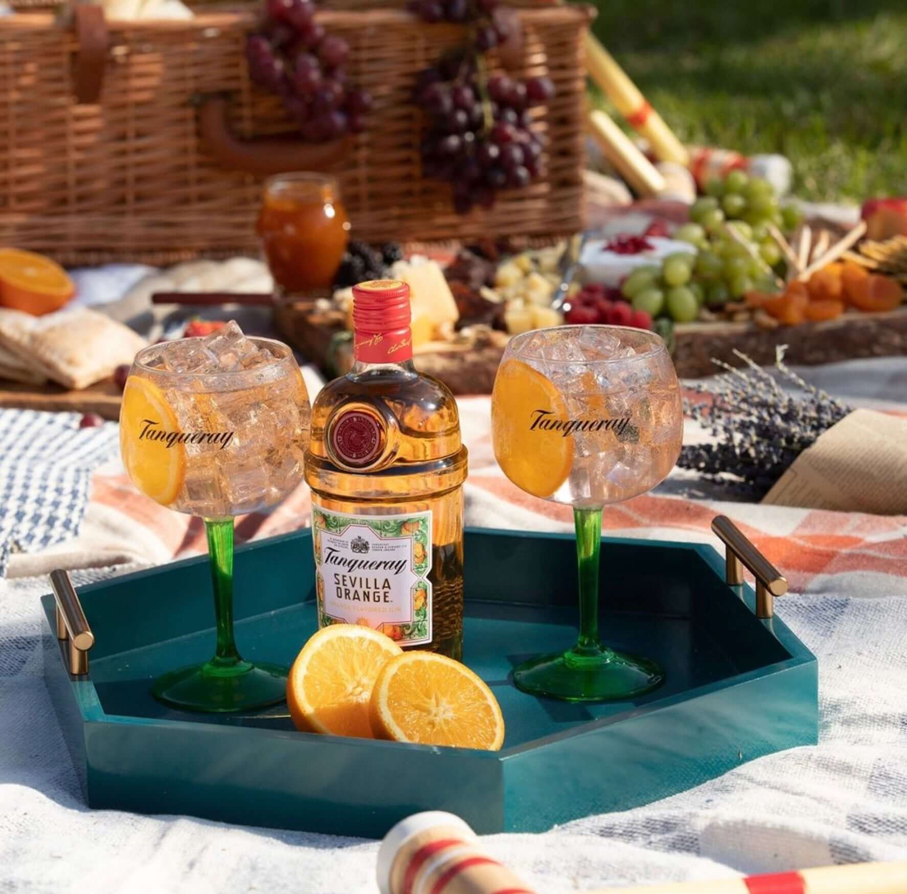 Tanqueray Sevilla Orange Gin on a picnic tray with orange slices and two glasses filled with ice, surrounded by a variety of snacks.