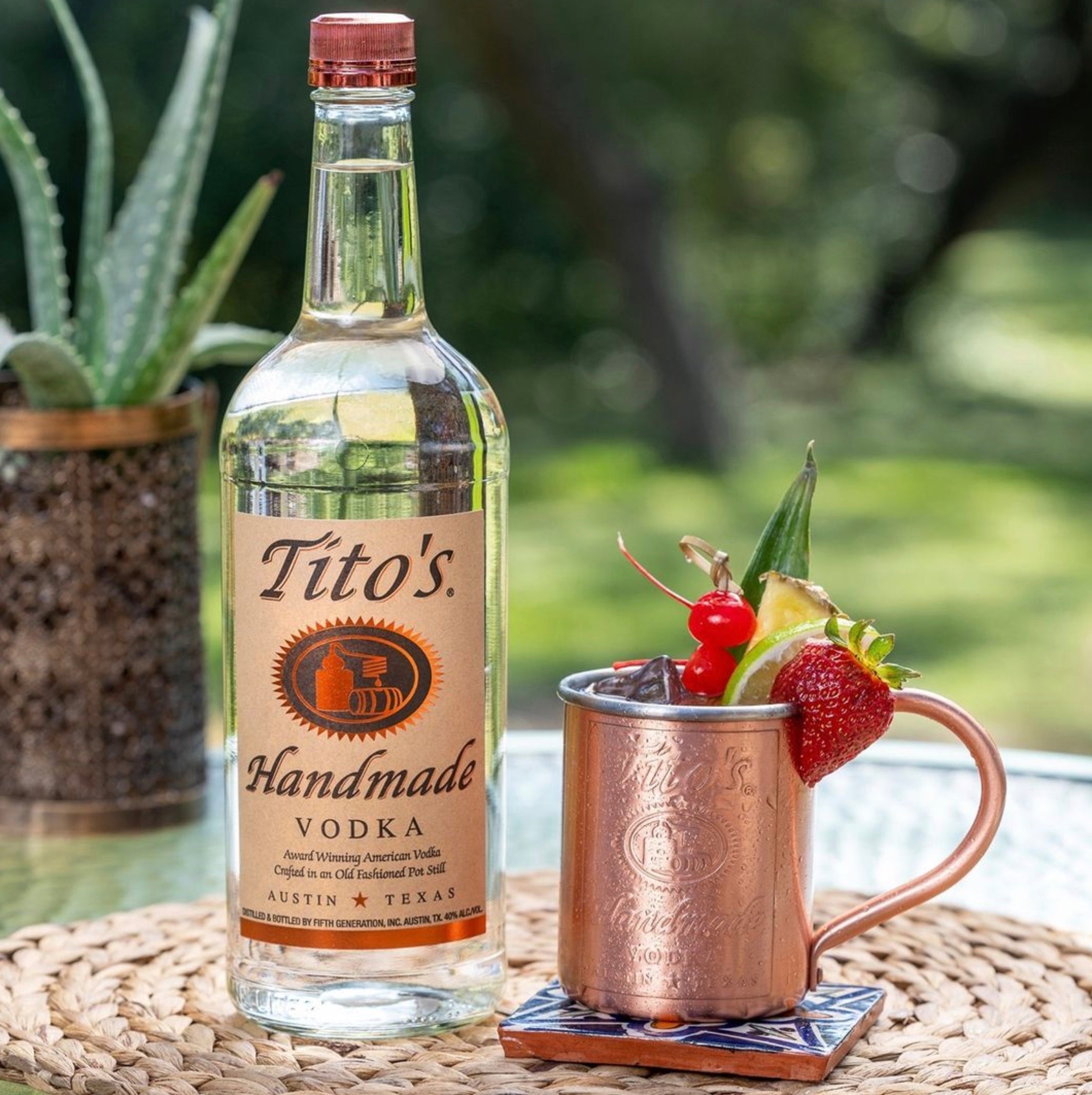 Bottle of Tito's Handmade Vodka next to a copper mule cocktail with garnishes, set in a lush outdoor scene.