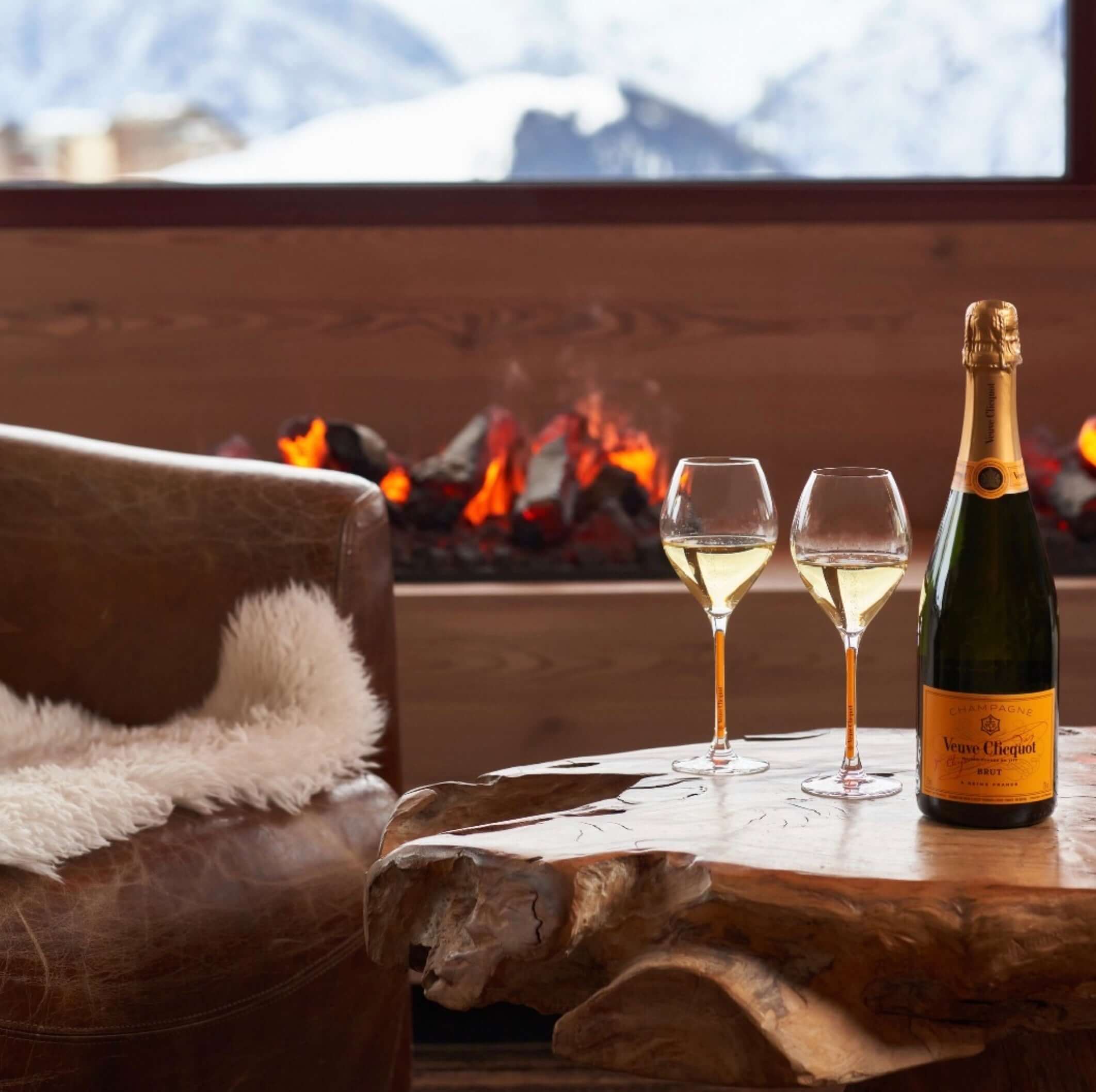 Veuve Clicquot Brut Champagne on a wooden table with two glasses, cozy setting by a fireplace and snowy mountains in the background.