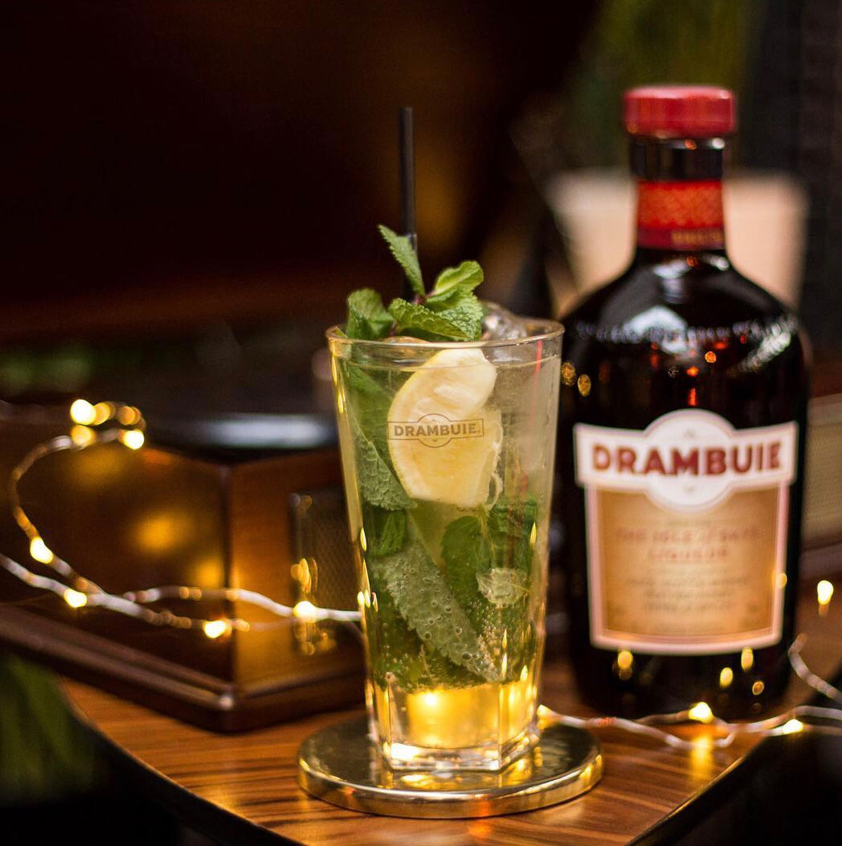 Refreshing Drambuie cocktail garnished with mint and lemon, alongside a bottle of Drambuie liqueur, set in a cozy atmosphere.