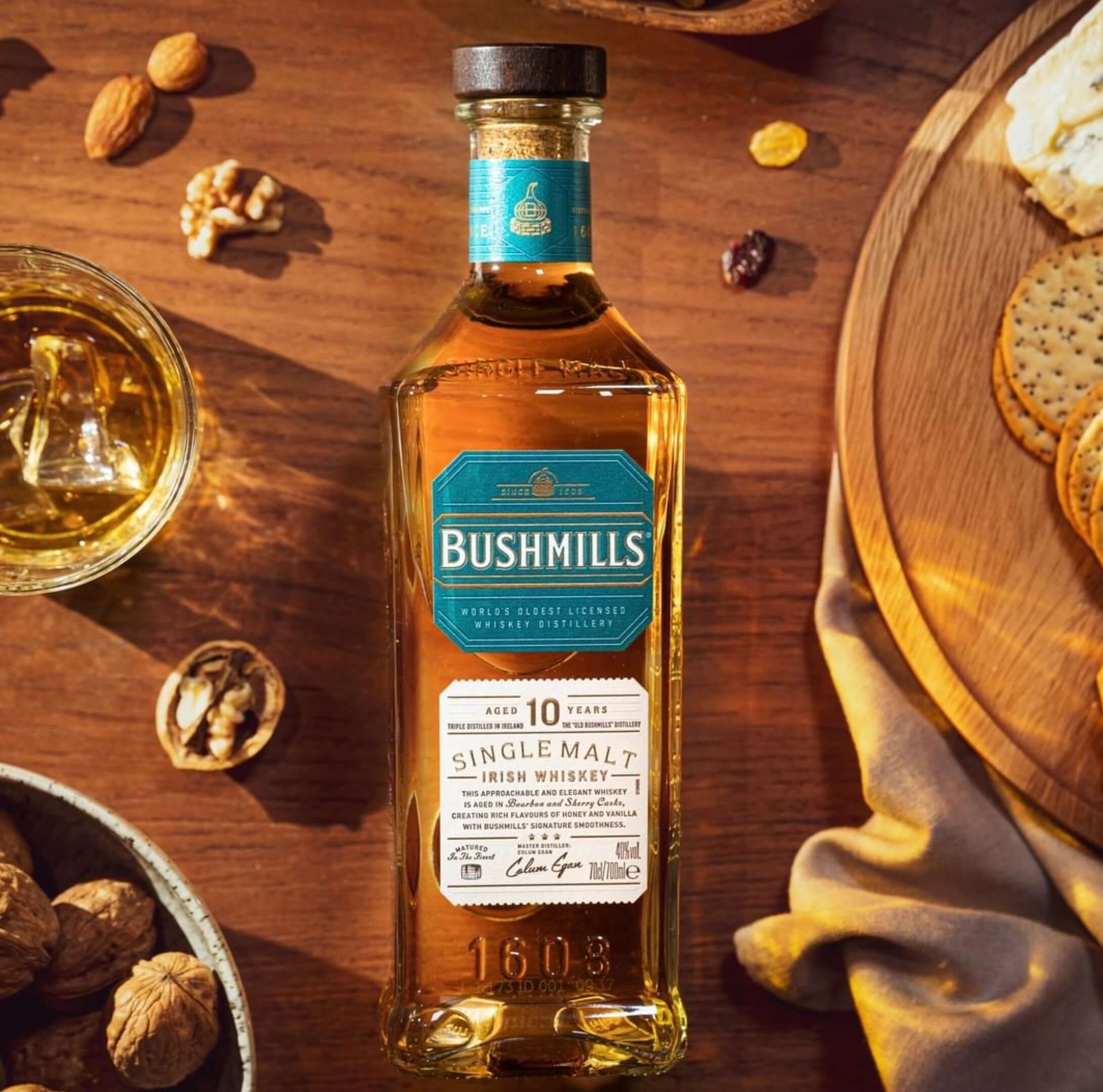 Bushmills 10 Year Single Malt Irish Whiskey bottle with glass, nuts, and crackers on a wooden table.