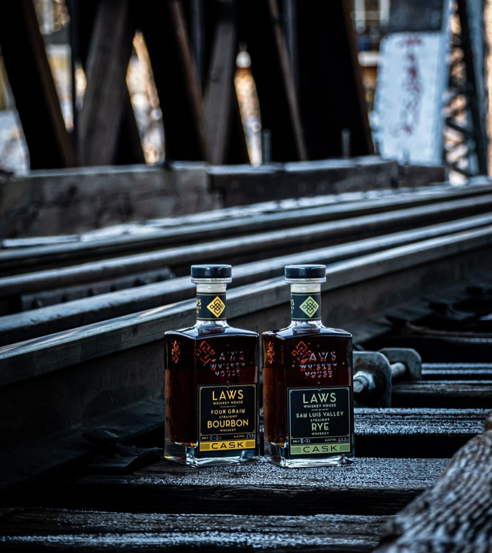 Laws Whiskey House Four Grain Bourbon and Rye displayed on railroad tracks, showcasing cask strength premium spirits.