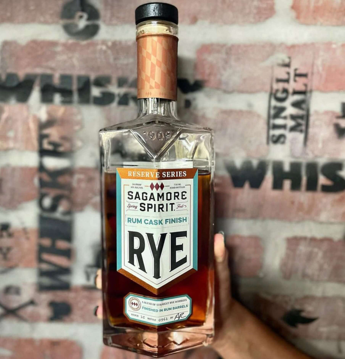 Sagamore Rye Rum Cask Finish Whiskey bottle featuring a decorative label, held against a brick wall.