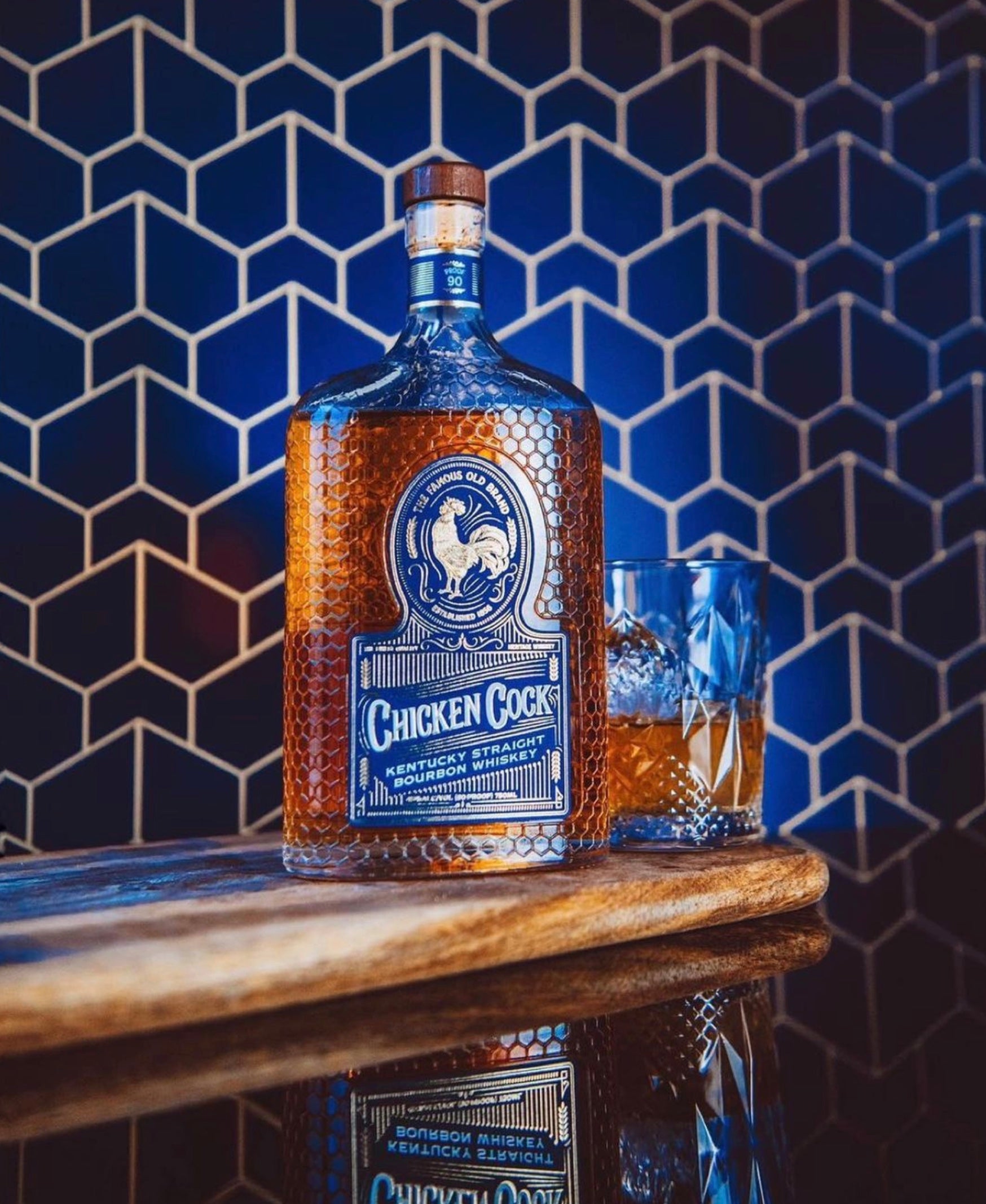 Bottle of Chicken Cock Kentucky Straight Bourbon with a glass, set against a stylish blue honeycomb background.