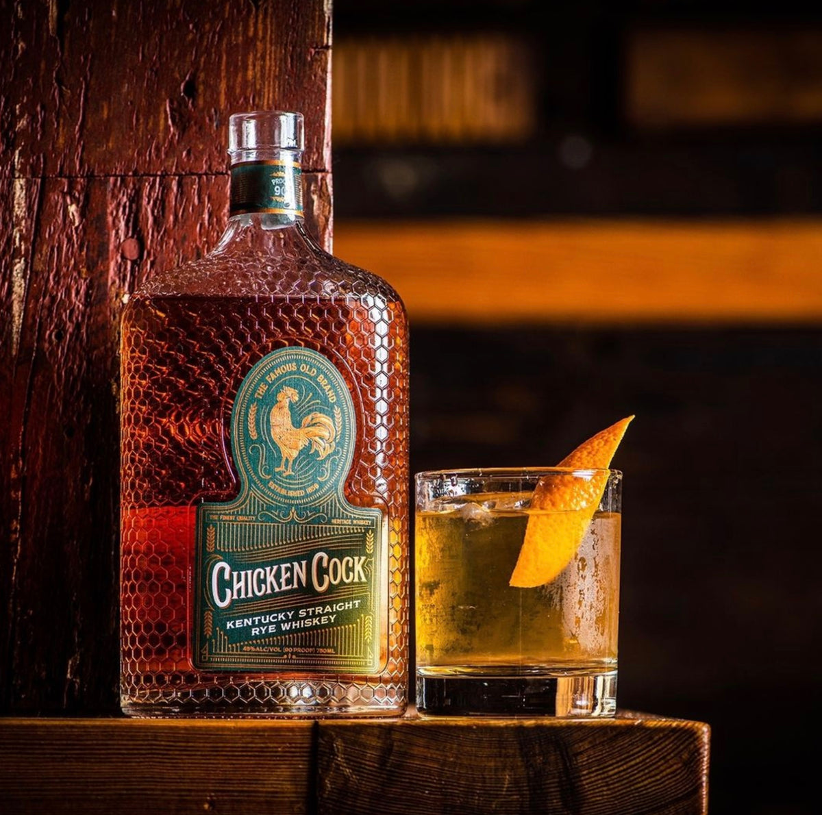 Chicken Cock Kentucky Straight Rye whiskey bottle next to a glass with cocktail and orange garnish.