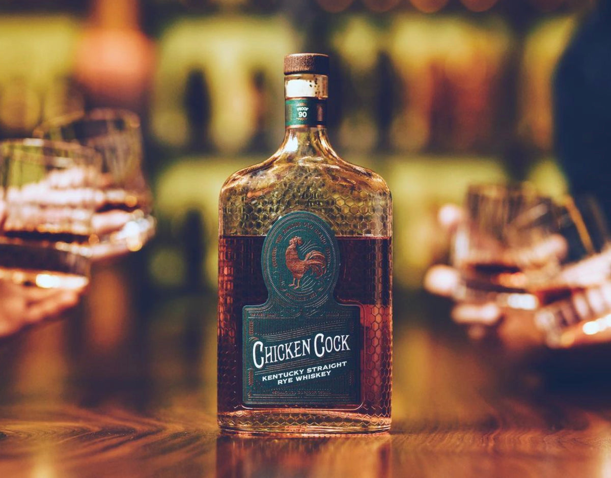 Bottle of Chicken Cock Kentucky Straight Rye whiskey surrounded by cocktail glasses on a wooden bar.